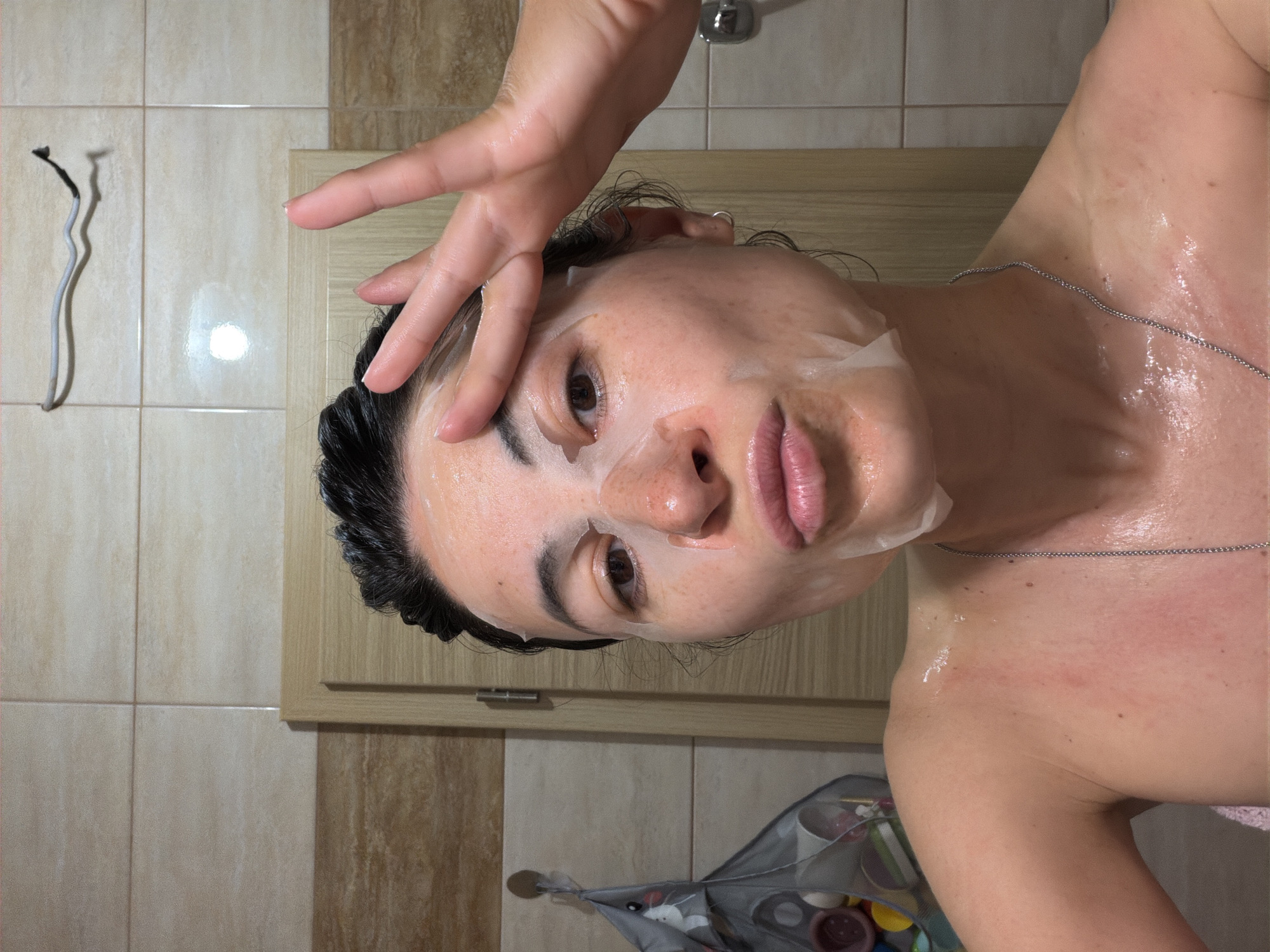 A woman with dark hair is wearing a sheet face mask and is in a bathroom. She is looking at the camera with a hand pressed against her forehead. She is wearing a silver necklace. The background consists of beige tile walls and a light wood cabinet. To the left of the frame is a drying rack with items in it. The location of the image is not discernible from the image alone. The city of Budva, Montenegro cannot be determined from the image.