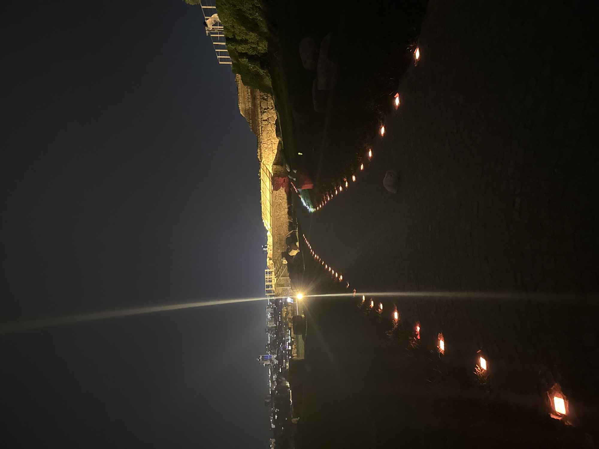 The image depicts a night scene in Plovdiv, Bulgaria. A stone wall with partial illumination runs vertically on the left side of the frame, separating a dark sky above and a cityscape with lights below. A pathway stretches from the lower right towards the left, lined with small, orange-glowing light fixtures. The pathway and the wall are bordered by dark greenery. People are dimly visible near the stone wall. The overall setting suggests a historical location at night with modern lighting.