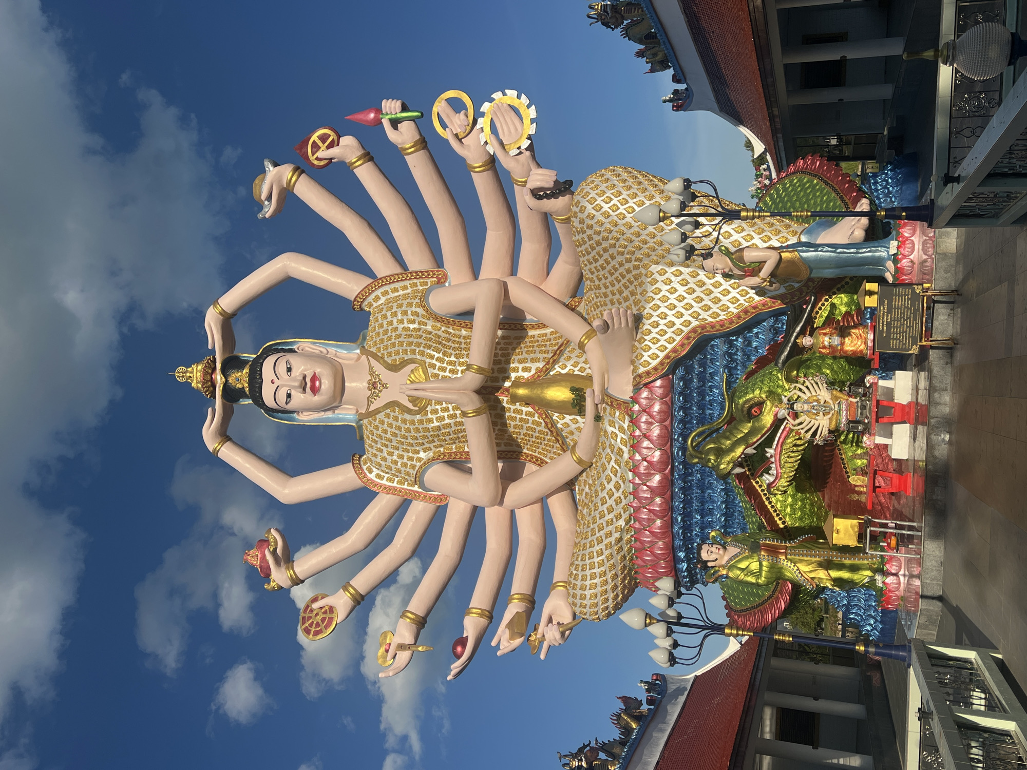 This media file captures a striking sculpture of a multi-armed deity, likely of Buddhist or Hindu inspiration, against a bright blue sky dotted with white clouds. The sculpture is adorned in a shimmering gold and white patterned robe and is depicted with numerous arms, each holding a symbolic object such as a wheel, a lotus, or a fruit. The deity's face is serene with painted features.
In the background, architectural elements of a temple or religious site are visible, featuring red tiled roofs and ornate details. A large, colorful dragon sculpture, predominantly green with red and blue accents, is positioned to the right of the main deity, coiled and appearing to guard the structure. Another smaller figure, dressed in green and gold, is seated near the dragon.
A lamppost with multiple white lights stands in the foreground, to the right of the frame. The scene is brightly lit, suggesting it is daytime, likely with clear weather. The overall impression is one of spiritual grandeur and intricate artistry. Some Thai text is visible on a sign in the lower right quadrant of the image, near the dragon sculpture. The location context identifies this as Baan Plai Laem, Thailand.