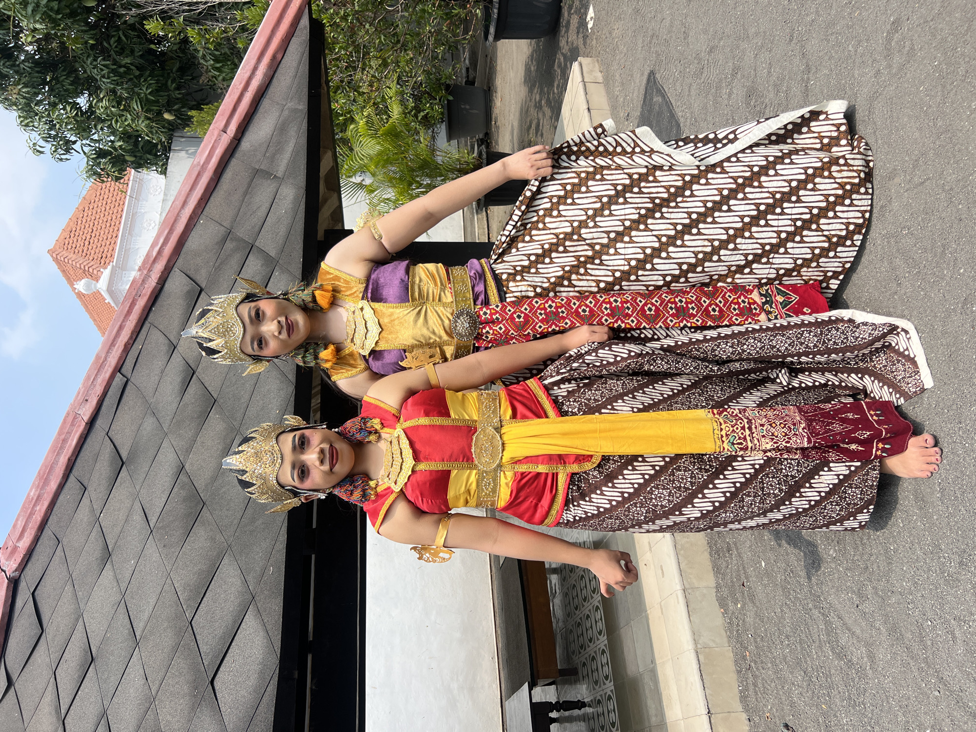 Two young women in traditional Javanese dance costumes stand outdoors in front of a building. The building has a dark, textured roof and a white facade with architectural details, including a red-tiled roof on a separate structure in the background. Lush green plants in black pots are visible to the right. The ground is paved with asphalt, with a tiled walkway in the foreground.
The woman on the left wears a red and yellow bodice, a gold belt, and a brown batik skirt with a white patterned motif. She has a gold headdress adorned with jewels and earrings. Her makeup is dramatic, with bold eye and lip color.
The woman on the right wears a gold and purple sleeveless top, a gold belt, and a batik skirt with a red and white geometric pattern. She also wears a gold headdress and earrings. Her makeup is similar to the woman on the left.
Both women are barefoot. They appear to be posing for a photograph. The overall impression is that they are performers at a cultural event or tourist attraction in Yogyakarta, Indonesia.