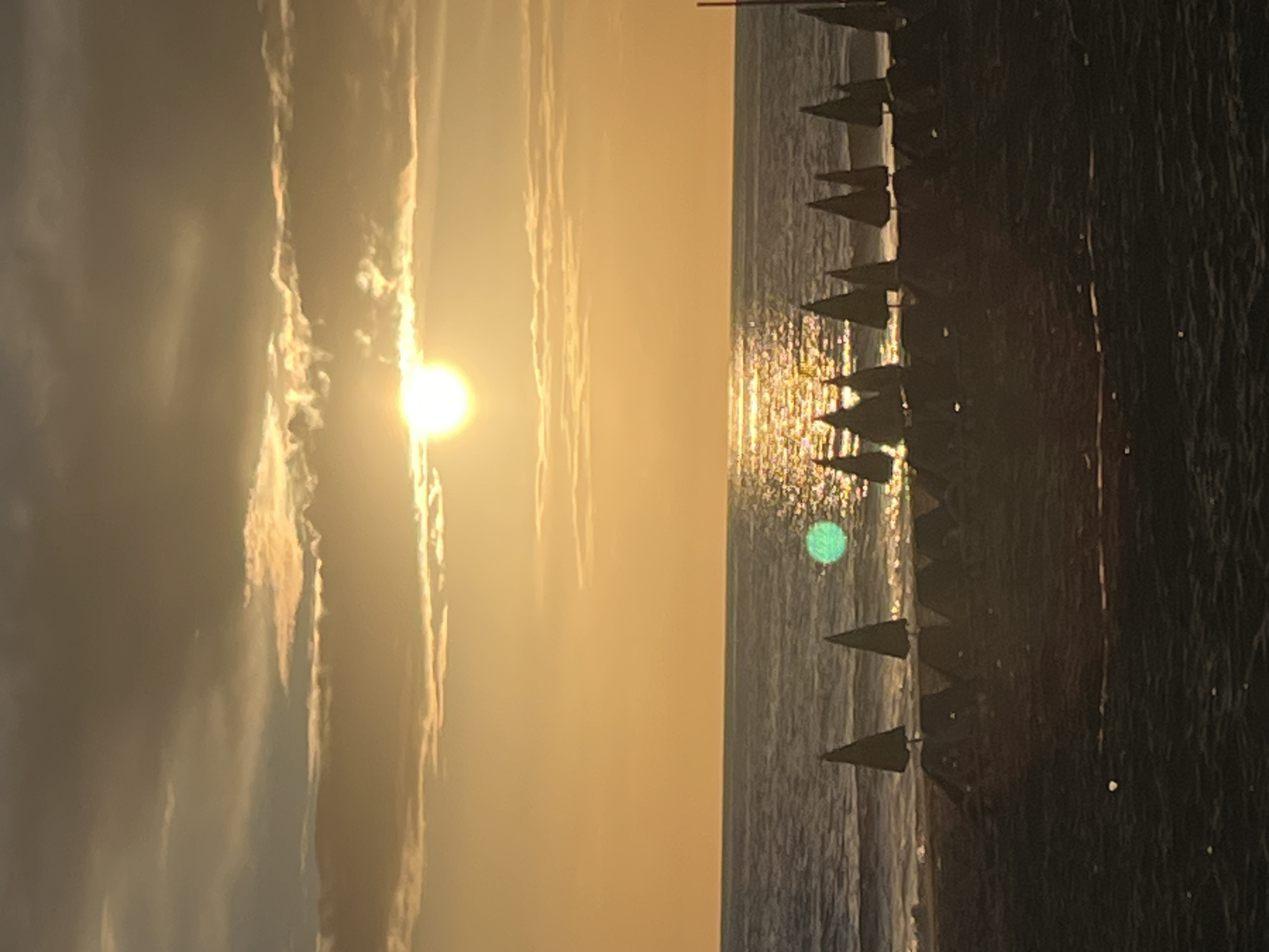 The image shows a sunset scene at a beach in Alanya, Turkey. The sun is positioned near the horizon on the left side of the frame, casting a golden light on the sky and clouds. The sea occupies the central portion of the frame, reflecting the sunlight. The right side of the frame features a row of sun umbrellas along the beach, silhouetted against the illuminated sea.