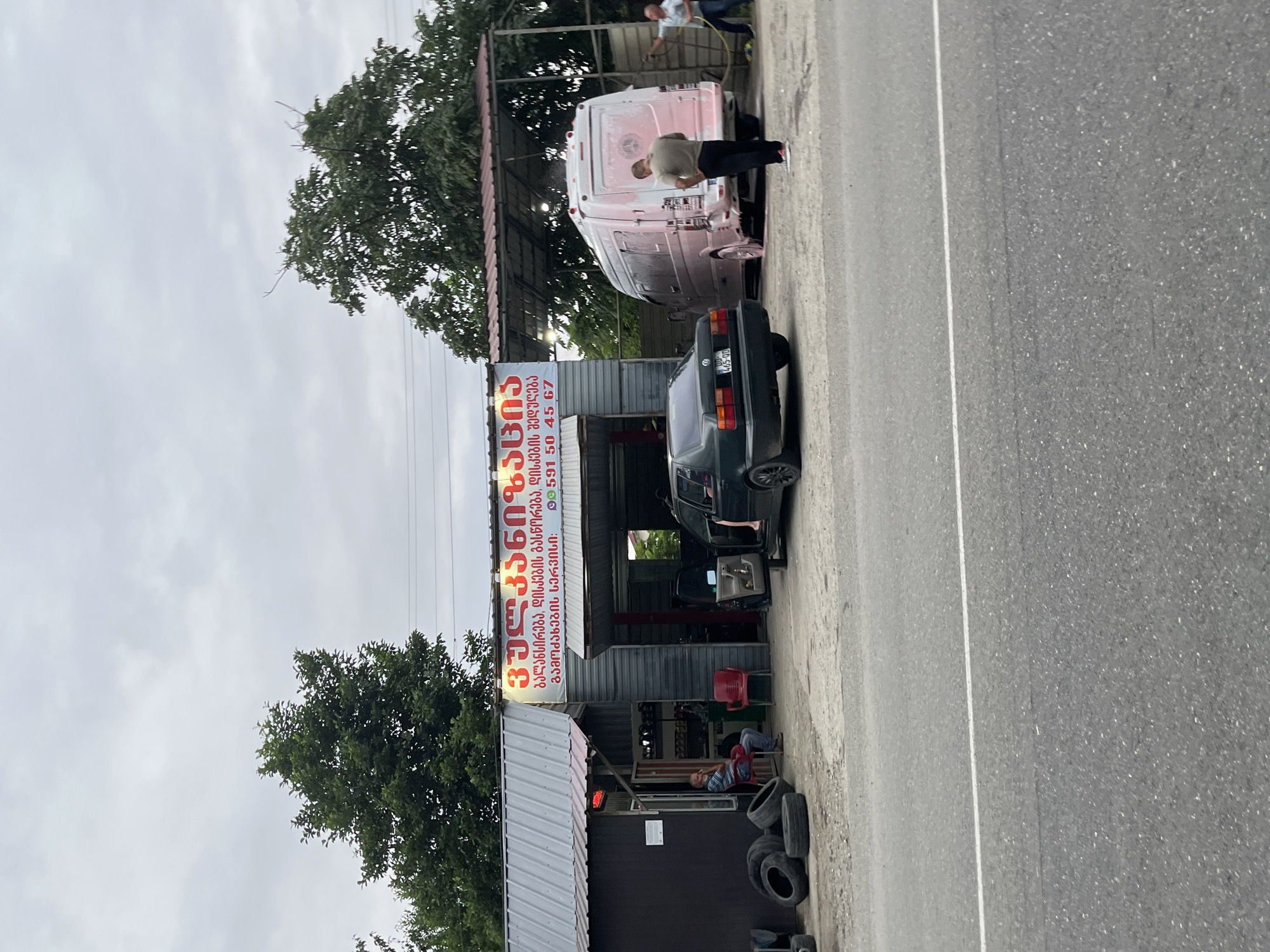 The image depicts a car wash service located in ღრმაღელის თემი, Georgia. A grey sedan is parked in front of an open garage, and a white van, partially covered in pink foam, is being washed by two men. One man is spraying the van with a hose, while another stands nearby observing. Several tires are stacked to the left of the service bay. The sky is overcast.