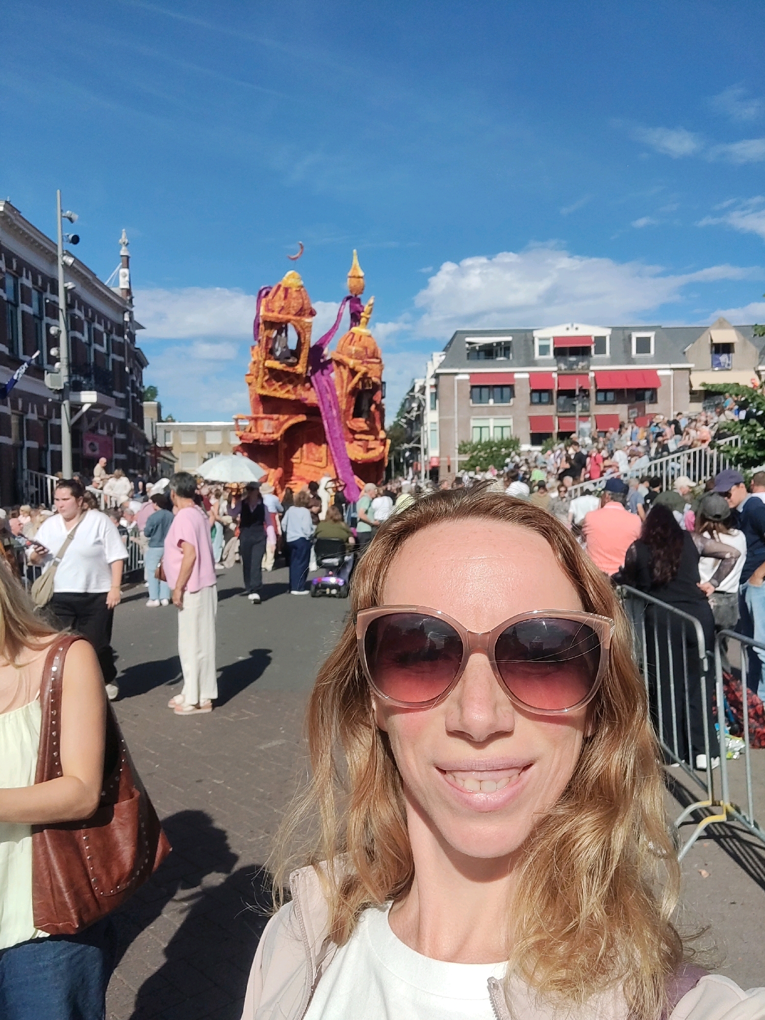 The image captures a bright, sunny day scene at the Zundert Flower Parade in Zundert, Netherlands. In the foreground, a woman with blonde, wavy hair and large pink sunglasses smiles directly at the camera, her face taking up a significant portion of the frame. She is wearing a light-colored jacket over a white shirt. To her left, another woman is partially visible, wearing a light green top and a brown purse with studded details.
The background reveals a street lined with buildings and a crowd of people. A large, elaborate float, decorated with orange and purple elements and resembling a castle or ornate structure, is the central focus of the parade. Spectators are gathered on both sides of the street, some standing, others sitting on risers, and some appear to be in a wheelchair. The street itself is paved with cobblestones, and a metal barrier separates the parade route from the onlookers. The sky is clear and blue with a few scattered clouds. The overall atmosphere suggests a lively outdoor event.