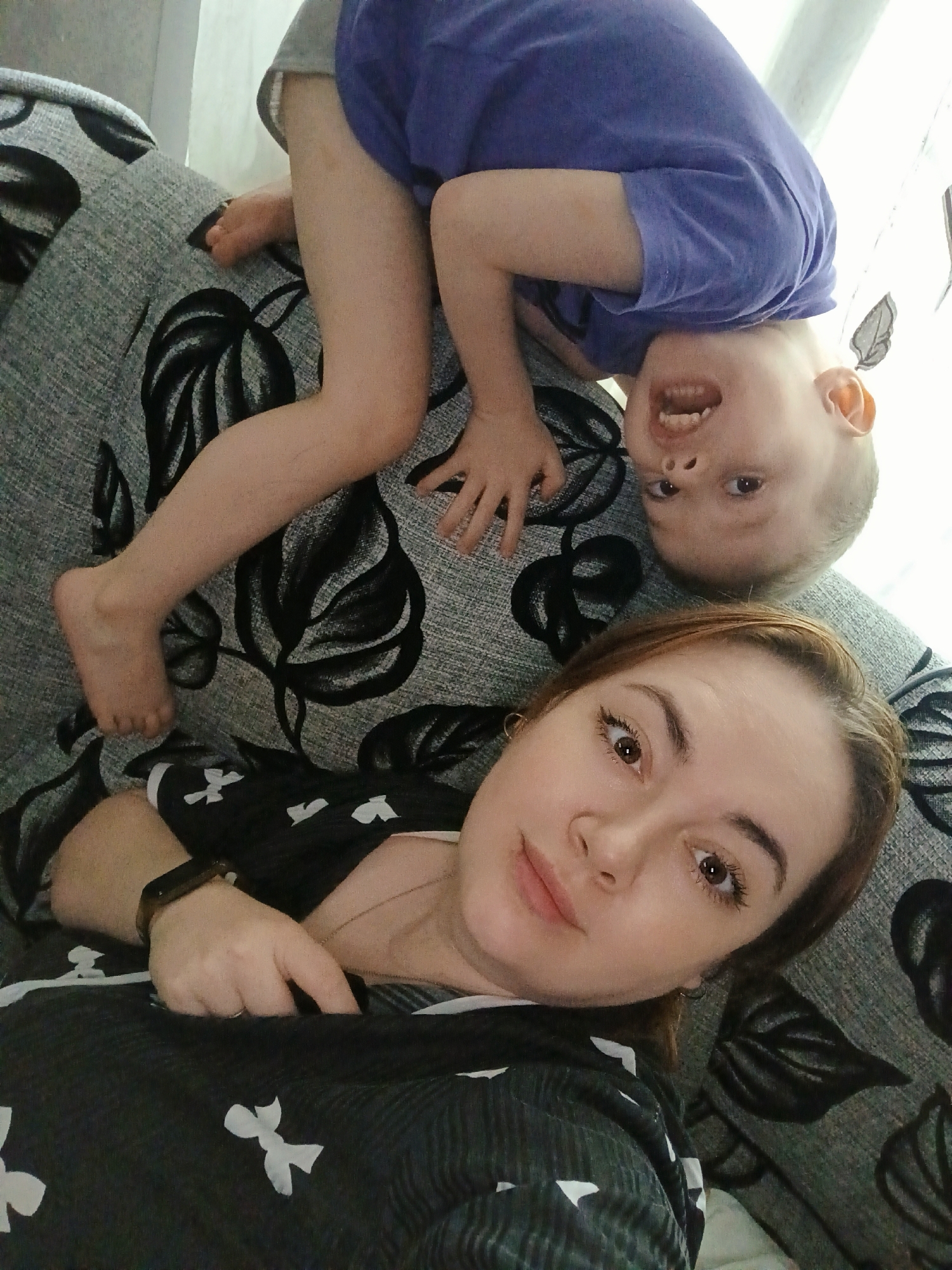A selfie captures a fair-skinned woman and a young child on a patterned couch, likely in a home setting. The woman is in the lower portion of the frame, facing the camera with a slight smile. She has brown hair and is wearing a black and white patterned top with a fitness tracker on her wrist. The child, dressed in a blue shirt and grey shorts, is positioned upside down above the woman. The child is also fair-skinned and smiles with their teeth visible. The couch has a grey background with black floral patterns. The location is Chaikovsky, Russia.