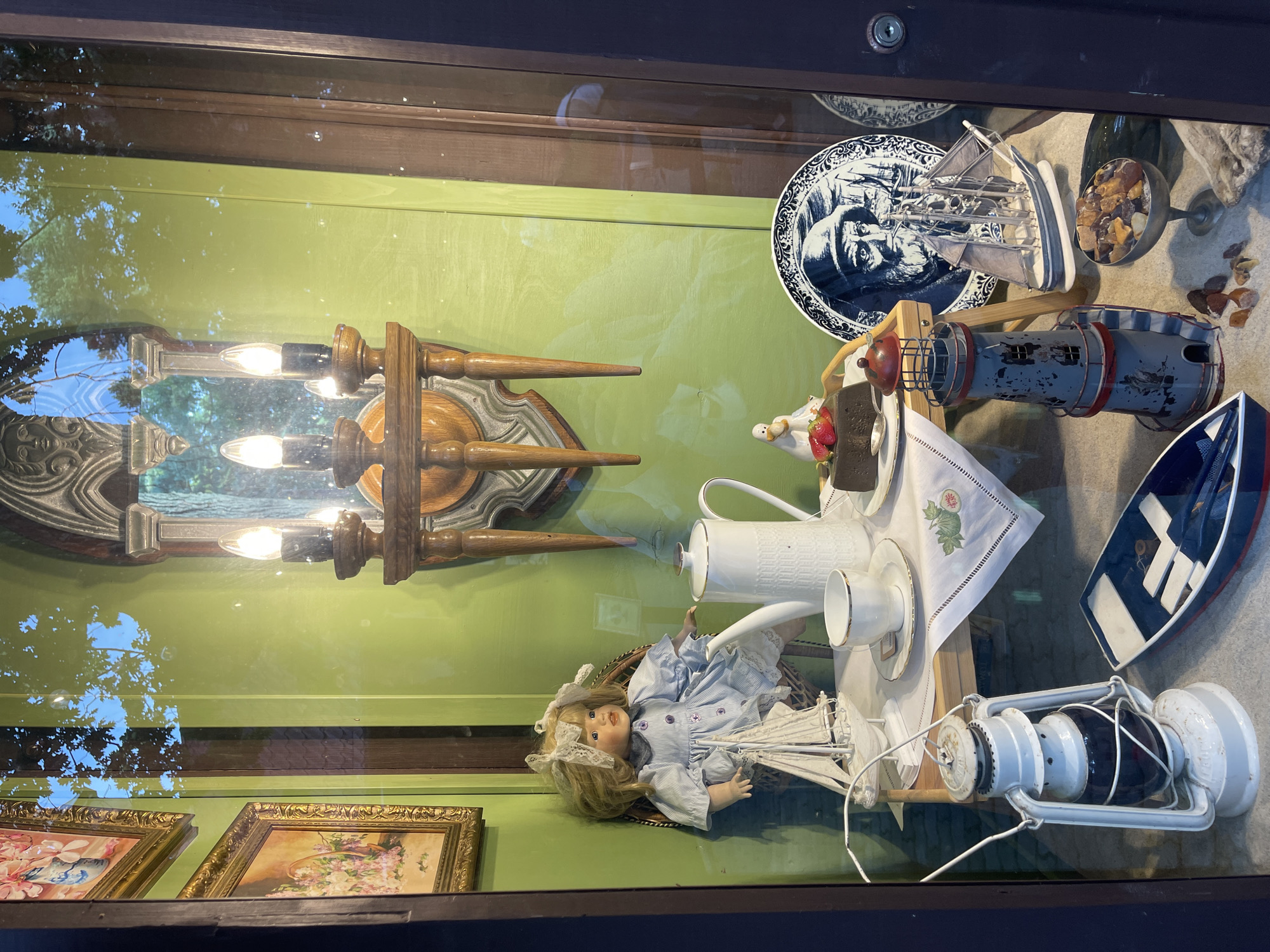 This image captures a display window in Svetlogorsk, Russia, featuring a collection of maritime-themed and vintage items. In the background, a bright green wall is adorned with an ornate, carved wooden sconce holding a three-bulb electric light fixture. To the left, two framed paintings depicting floral arrangements hang on the wall. The central focus is a small table set for tea. A porcelain doll with blonde hair, wearing a blue dress and white bow, sits in a wicker chair. Nearby, a white teapot, a teacup and saucer, and a small cake topped with strawberries are arranged on a white tablecloth. A miniature white sailboat rests on the table.
Below the table, several other objects are displayed on what appears to be a sand-covered surface. These include a white oil lantern, a small blue and red model boat, a detailed model lighthouse, and a decorative plate featuring a portrait of a man with a beard and hat. A miniature sailboat is also positioned near the plate. In a small silver goblet, amber-like colored stones are visible. The entire scene is framed by reflections of trees and sky on the glass of the display window, indicating an outdoor setting.