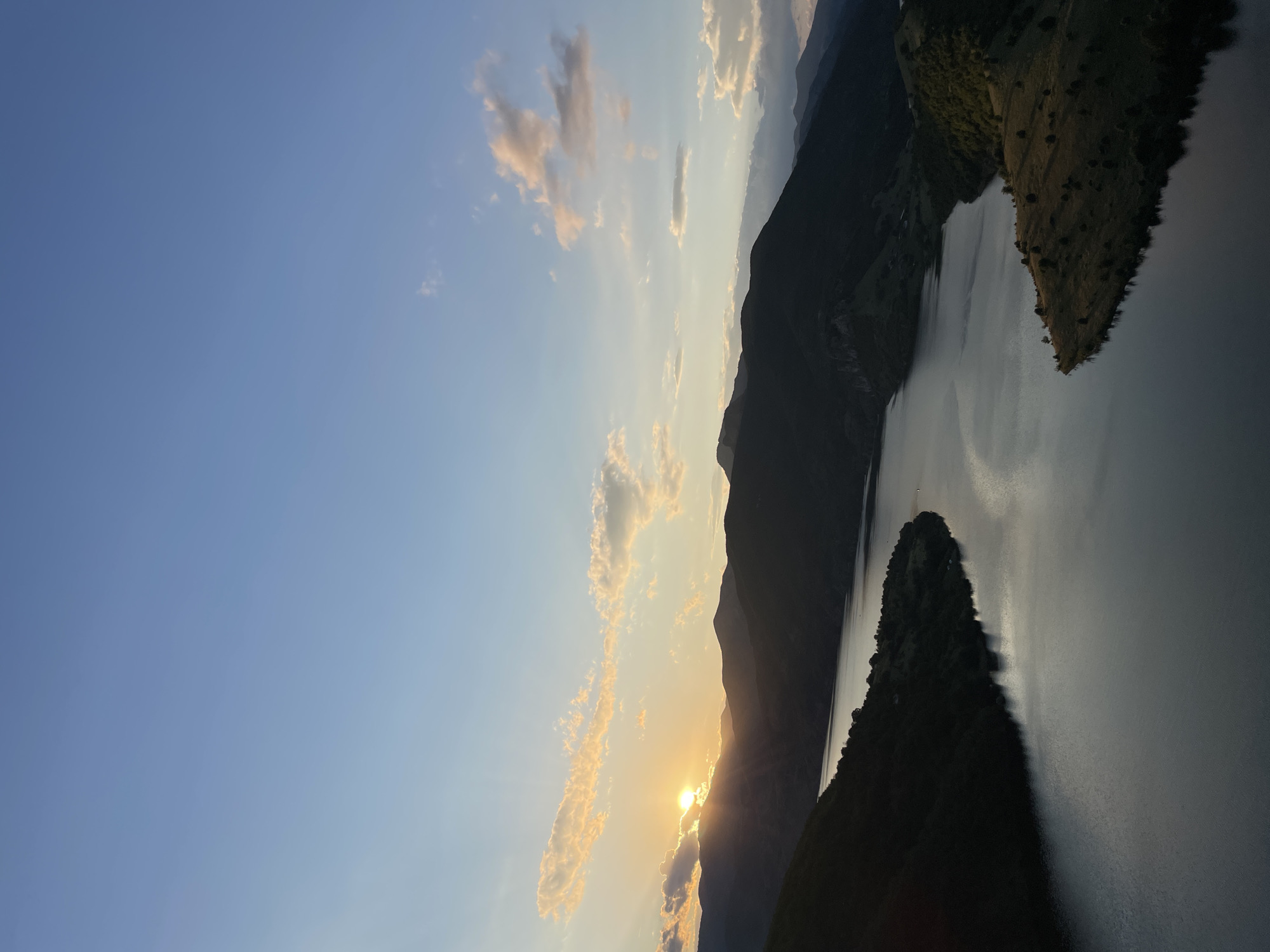 An aerial, high-angle, long shot captures a serene sunset over a vast body of water, likely a reservoir or lake, nestled amidst a chain of dark, silhouetted mountains in Borovitsa, Bulgaria. The sun, a bright orb peeking from behind a jagged mountain peak on the left, casts a warm, golden glow, illuminating scattered, wispy clouds with hues of orange and pink. The tranquil water reflects the soft light of the sky, creating a shimmering, almost metallic surface. The surrounding landscape is a study in contrasts: the rugged, dark mountains frame the luminous sky and water, while the foreground shows rolling hills dotted with sparse trees, bathed in the fading sunlight. The scene is devoid of human presence, emphasizing the raw beauty and solitude of the natural environment. The sky transitions from a pale blue near the horizon to a deeper shade of cerulean overhead. The image evokes a sense of peace and natural grandeur.