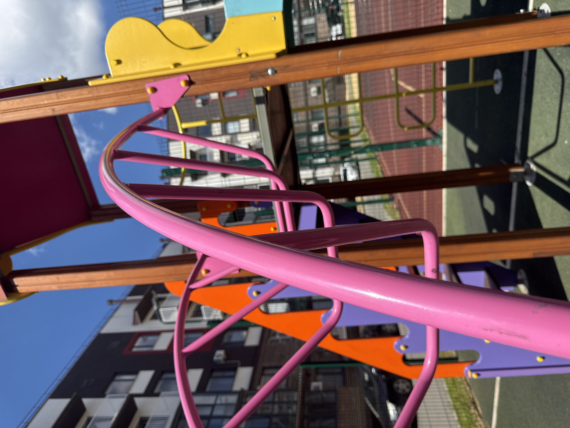 A close-up, low-angle shot of a brightly colored playground structure, with pink, orange, and yellow elements prominently featured. The playground is situated in a modern urban setting, with contemporary apartment buildings visible in the background. The sky is clear and blue with scattered white clouds. The ground surface appears to be a mix of green rubberized material and red track-like flooring, common in public play areas. The image captures a sunny day, with shadows cast by the playground equipment. The location is identified as Vyazniki, Russia.
