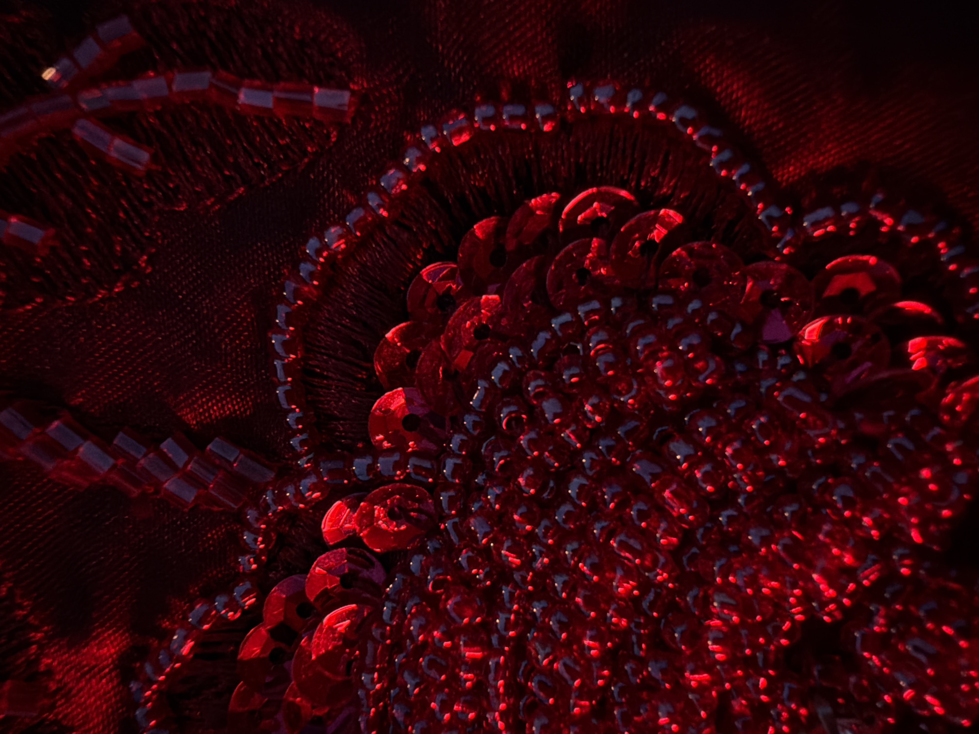 The image shows a close-up, abstract view of red fabric adorned with sequins and beads. The lighting is dramatic, casting deep shadows and highlighting the reflective surfaces of the embellishments. The texture of the fabric appears to be a fine mesh, overlaid with a denser, slightly textured material. The sequins are circular and faceted, catching the light to create bright red highlights. Interspersed among the sequins are small, rectangular beads that are darker in tone, appearing almost black in the low light. The arrangement of the embellishments suggests a floral or intricate decorative pattern, though the full design is not visible due to the close-up perspective. The overall impression is one of opulent texture and vibrant, saturated color. The scene is not identifiable as being in any specific city or location.