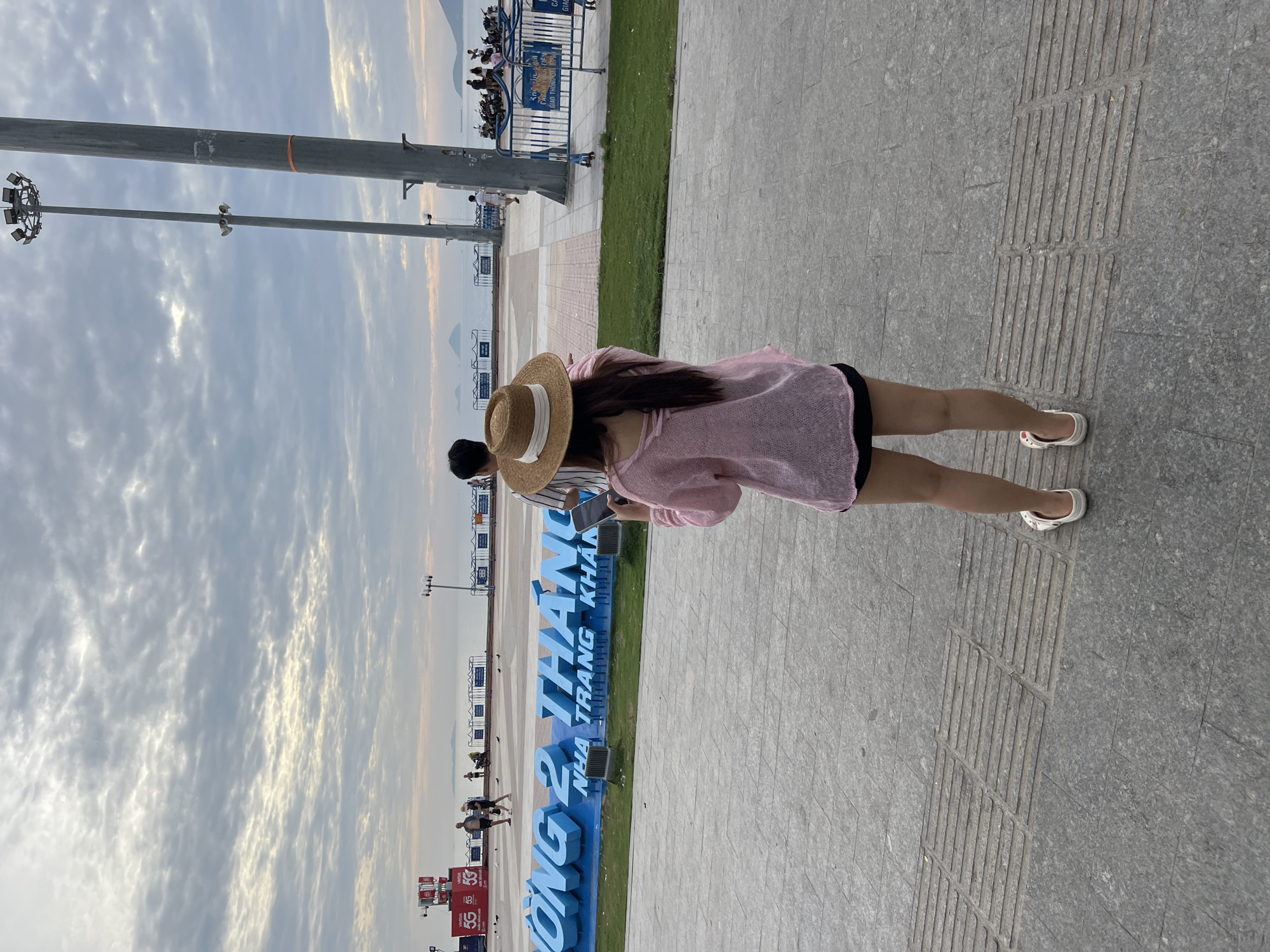 A rear view of a woman wearing a straw hat, a pink knitted sweater, and black shorts, holding a phone. She stands on a grey tiled walkway in front of a large, blue, 3D sign that reads "NG 2 THÁNG" on top and "NHA TRANG KHAN" below. Behind her, a man in a striped shirt stands next to her, also looking away. In the background, the ocean is visible with a cloudy sky above. Several people are walking and standing near the water, and some motorcycles are parked. Tall light poles stand on the right side of the image. The scene appears to be a public area in Nha Trang, Vietnam, likely a waterfront promenade.