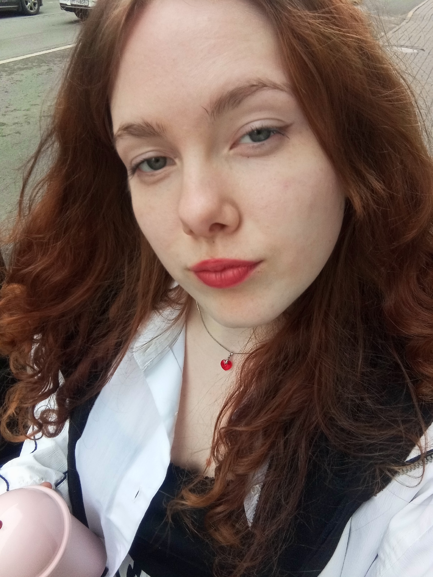 A reddish-haired young woman is taking a selfie outdoors. She is wearing a white shirt over a black top and has a heart-shaped pendant necklace. Her makeup includes red lipstick and eyeliner. She is holding a pink container in her left hand.
Based on context clues such as the user's location, and the architecture and road markings visible in the blurred background, the location is likely Saint Petersburg, Russia. However, specific details about the environment in the background are not clearly visible.
