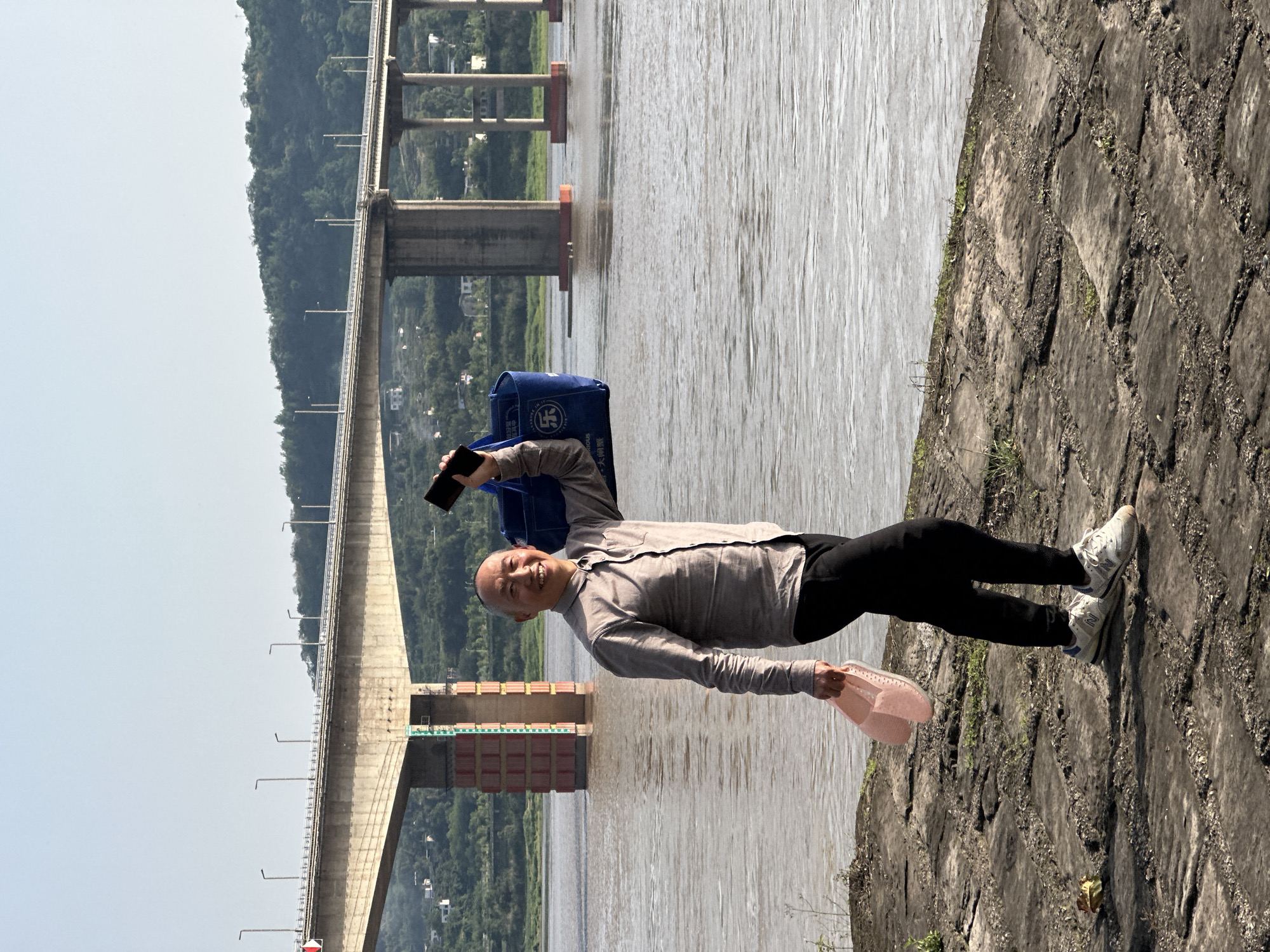 An older man poses for a photo near the Yangtze River in Jiangjin, China. The man, with short gray hair and wearing a gray long-sleeved shirt, black pants, and white sneakers, holds a smartphone in his left hand and pink shoes in his right. A blue bag rests on his left shoulder. In the background is the Yangtze River, spanned by a large bridge supported by concrete pillars. The immediate foreground consists of a gray stone embankment. Vegetation is interspersed on the hillside behind the bridge. The sky is overcast.