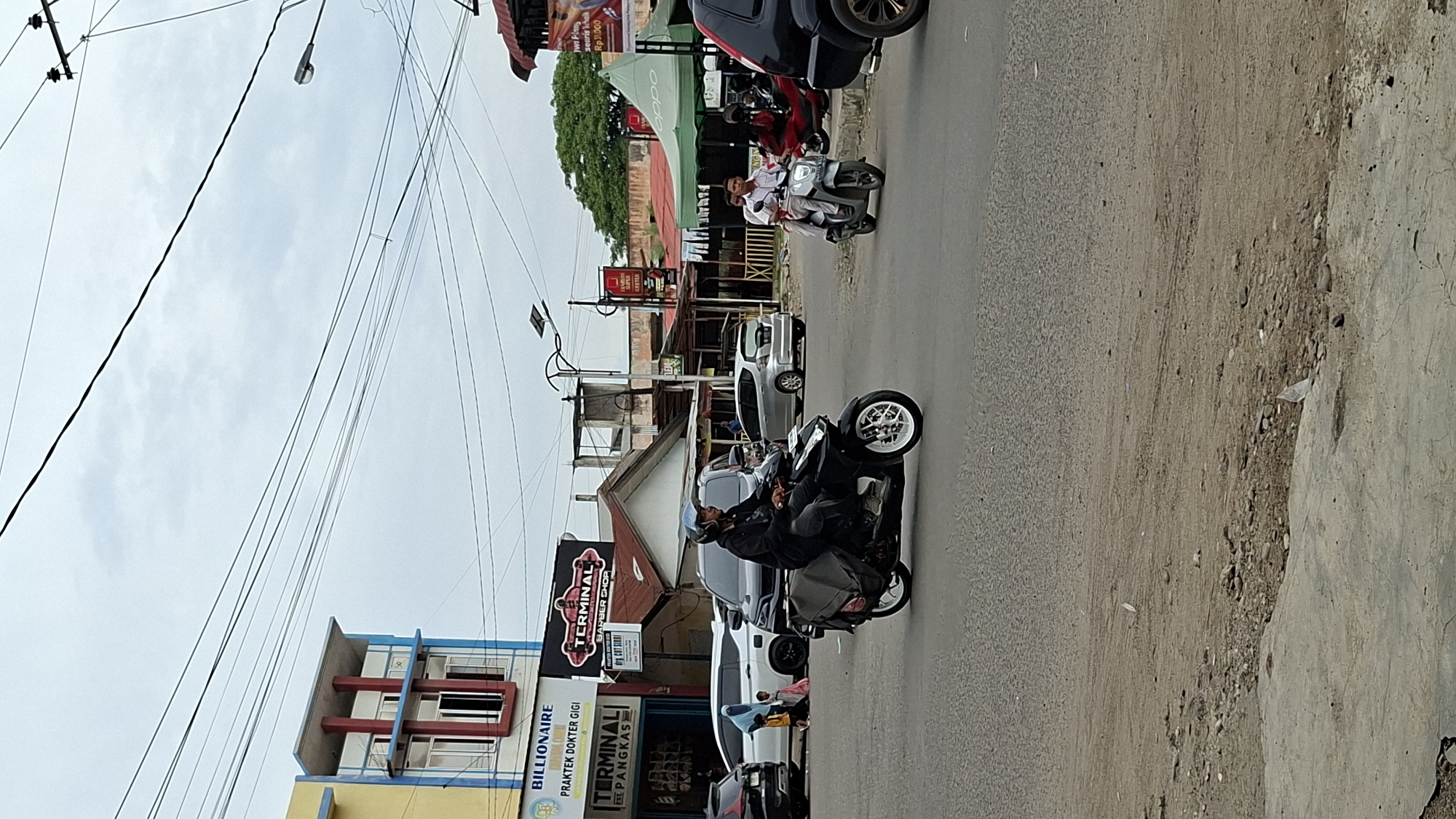 The image shows a street scene in Lhokseumawe, Indonesia. In the foreground, a man on a black scooter with white rims is riding on the asphalt road. He is wearing a dark jacket and a helmet. The scooter has saddlebags attached to the rear. In the background, several cars are parked and driving on the road. To the left, there is a building with yellow and blue accents, and a sign that says "BILLIONAIRE PRAKTEK DOKTER GIGI TERMINAL". Further back, there are other buildings, some with red roofs, and green awnings. A man and a woman are riding on a silver scooter in the mid-ground. Overhead, power lines crisscross the cloudy sky. The overall environment appears to be an urban or semi-urban area with commercial establishments.