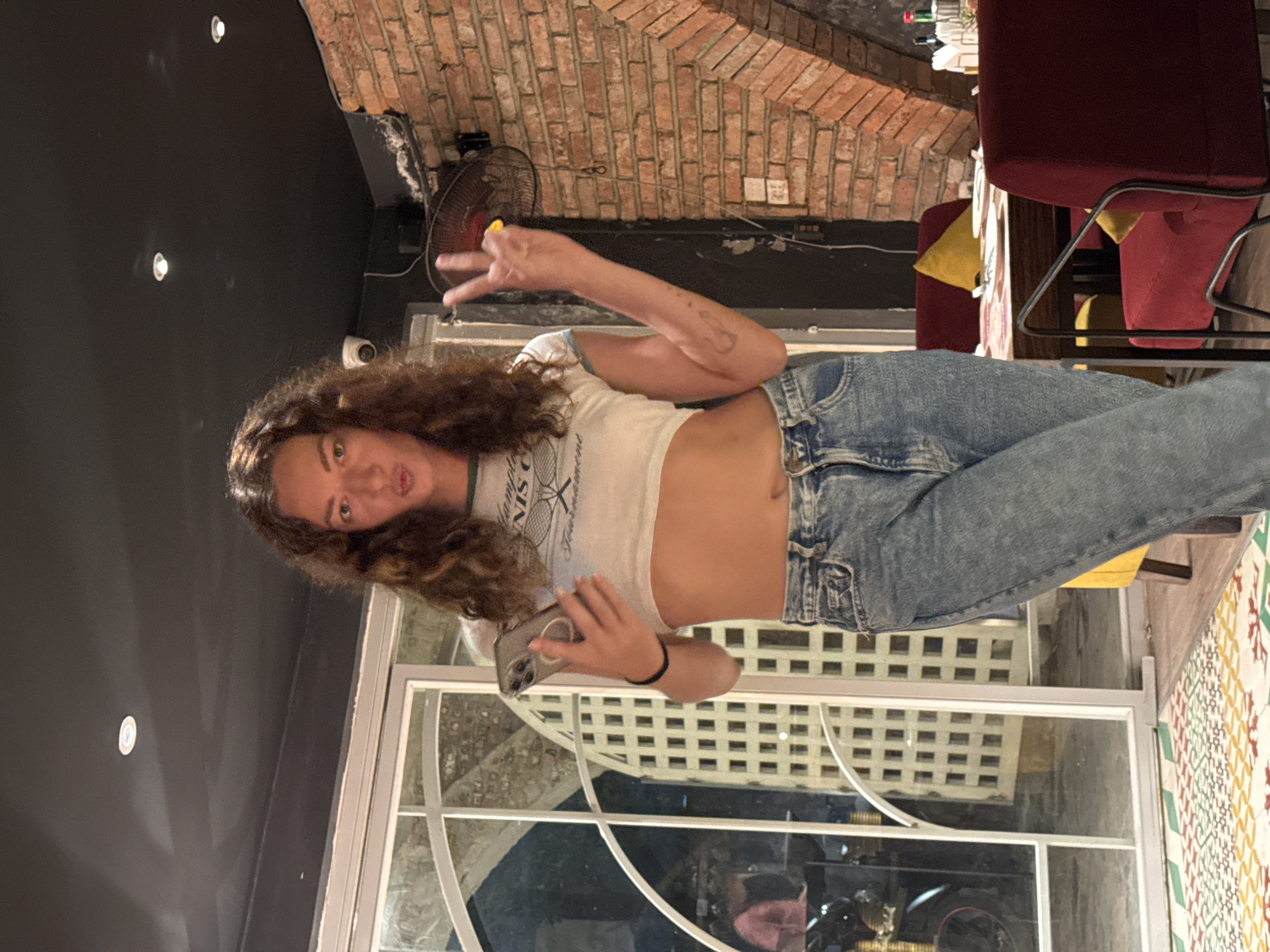 A young woman with curly brown hair stands in a dimly lit restaurant, holding a smartphone in her left hand and making a peace sign with her right hand. She is wearing a white crop top with a green trim and blue denim jeans. The background features a brick wall, a large glass-paned door, and a ceiling with lights. The floor has a colorful patterned rug. The woman is looking towards the camera with a playful expression. This scene appears to be in Hanoi, Vietnam, suggested by the presence of what looks like a Vietnamese street food stall reflected in the glass door and the style of the restaurant interior which often features exposed brick and intimate lighting in urban areas of Hanoi.
