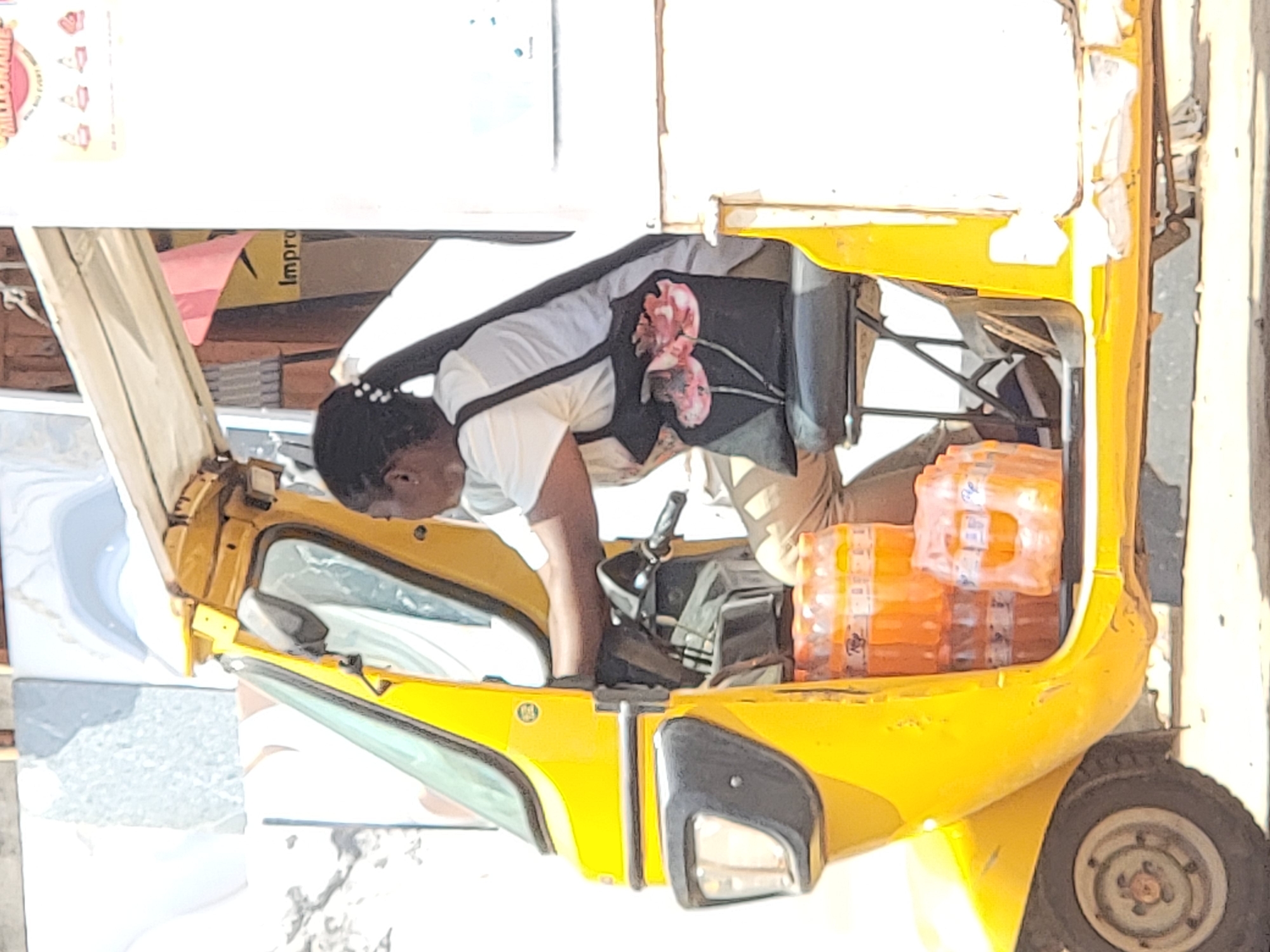 A woman leans into the front of a yellow, three-wheeled vehicle in Maiburiji, Nigeria. The vehicle is loaded with cases of bottled drinks. The woman has dark hair and wears a white shirt, light-colored pants, and a black apron with a floral print. The vehicle is parked next to a stack of tiles and cardboard boxes, suggesting a commercial area. The lighting indicates a sunny day.