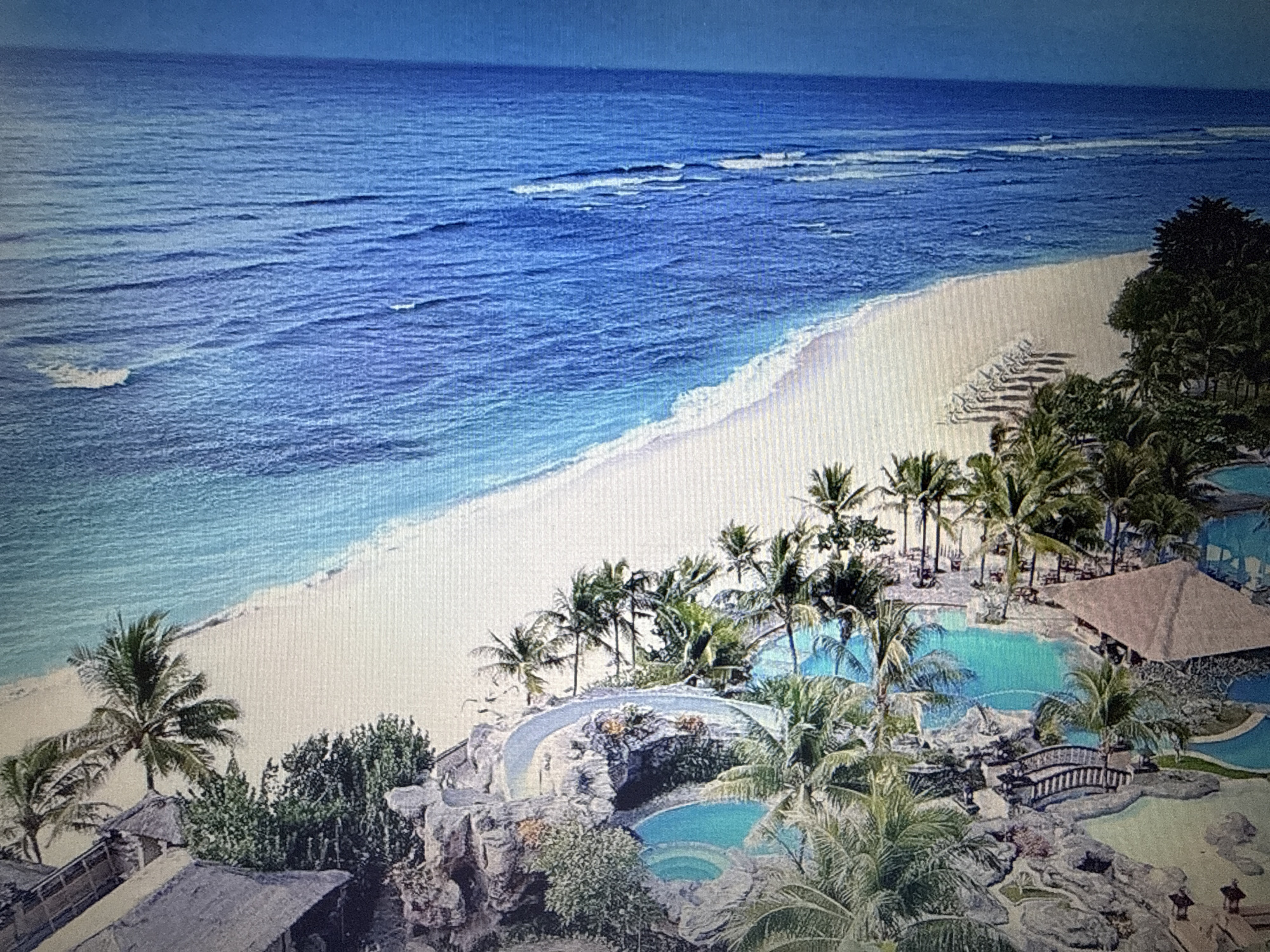 This is a high-angle, full shot of a tropical resort with a beach and ocean. The ocean is a deep blue with lighter turquoise shallows near the shore. Gentle waves break on a wide, white sand beach lined with lounge chairs. Lush green palm trees and other tropical foliage are abundant throughout the resort, which features multiple swimming pools, rock formations, and thatched-roof structures. A small, arched bridge crosses over one of the pools. The overall scene depicts a serene and luxurious vacation destination. The image is not located in Pushkino, Russia, but rather in a tropical coastal region.
