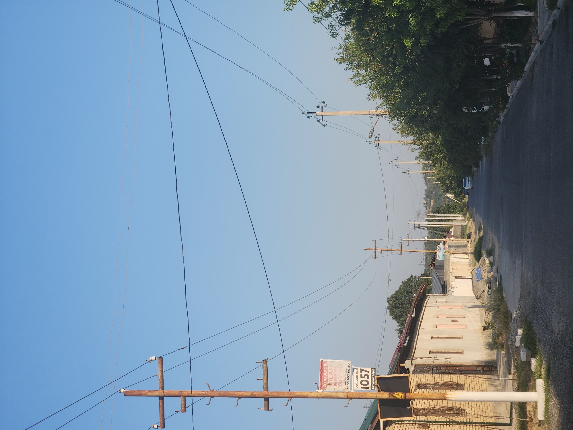 This image depicts a residential street in Xidirsho, Uzbekistan, under a clear blue sky. A prominent rusty metal utility pole stands on the left side of the road, adorned with a sign that reads "ABU AYUB GILAM MEBEL YUVISH" and a phone number, followed by "1057 bekijaan taxi". Adjacent to the pole is a building constructed with light-colored brick. Across the street, a row of trees lines the pavement, some with whitewashed trunks. Further down the street, the road curves gently, with more utility poles and trees visible. A white car is parked on the right side of the road, and another vehicle is moving away in the distance. The overall impression is one of a quiet, sunny day in a semi-rural or suburban setting.