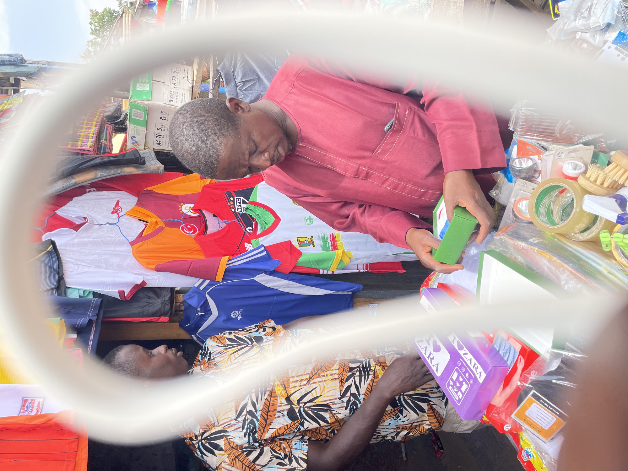 A medium shot, captured through an unclear lens (likely glasses), depicts a bustling open-air market scene in Jos, Nigeria. Two adult men are prominently featured: one is dressed in a maroon tunic, examining a small, green, rectangular object, while the second man, clad in a vibrant, patterned shirt, appears to be reaching for a purple box labeled "I <heart> ZARA".
Assorted objects surround the men, including various articles of clothing (primarily sports jerseys from different teams), office supplies such as tape and paper, cleaning supplies, and packaged items. Boxes, some labeled "A4 70 5", are visible in the background. The scene is outdoors, and overhead structures suggest a market stall or covered area.