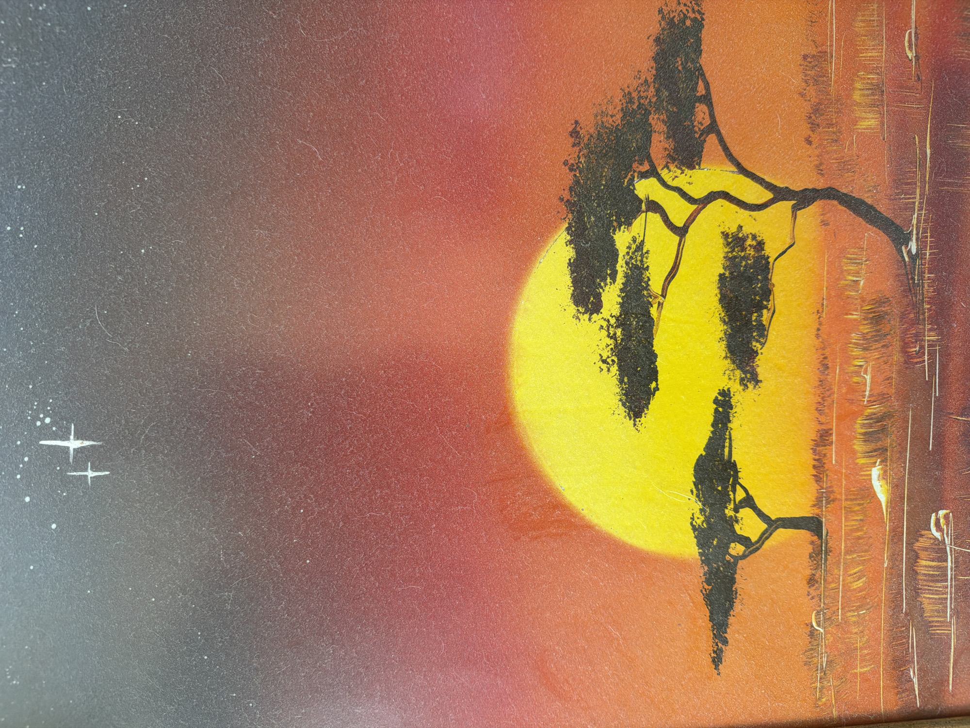An abstract and vibrant painting depicts a stylized sunset or sunrise scene. The left side of the composition features a dark gray-blue background, transitioning smoothly through a reddish-purple hue to a bright orange on the right. Small white specks, resembling distant stars, are scattered across the darker areas, with two more prominent, stylized white star shapes positioned in the upper-left quadrant.
A large, luminous yellow circular form, representing the sun or moon, dominates the right-center of the painting, casting a bright glow. Silhouetted against this intense yellow and the warm orange sky are the dark, intricate branches and foliage of what appear to be acacia or savanna trees. The trees are rendered in stark black, with a textured, almost speckled appearance, suggesting dense leaves or rough bark. Some branches extend horizontally across the celestial body, creating a striking contrast.
To the far right, the background shifts to deeper orange and brown tones, featuring vertical brushstrokes or textural elements that could represent distant trees, reflections, or an abstract textured surface. The painting exhibits visible surface texture, possibly from the canvas or painting technique (e.g., spray paint), and some scattered dust or light debris across its surface, which is more noticeable in the darker areas. No visible text is present in the image.