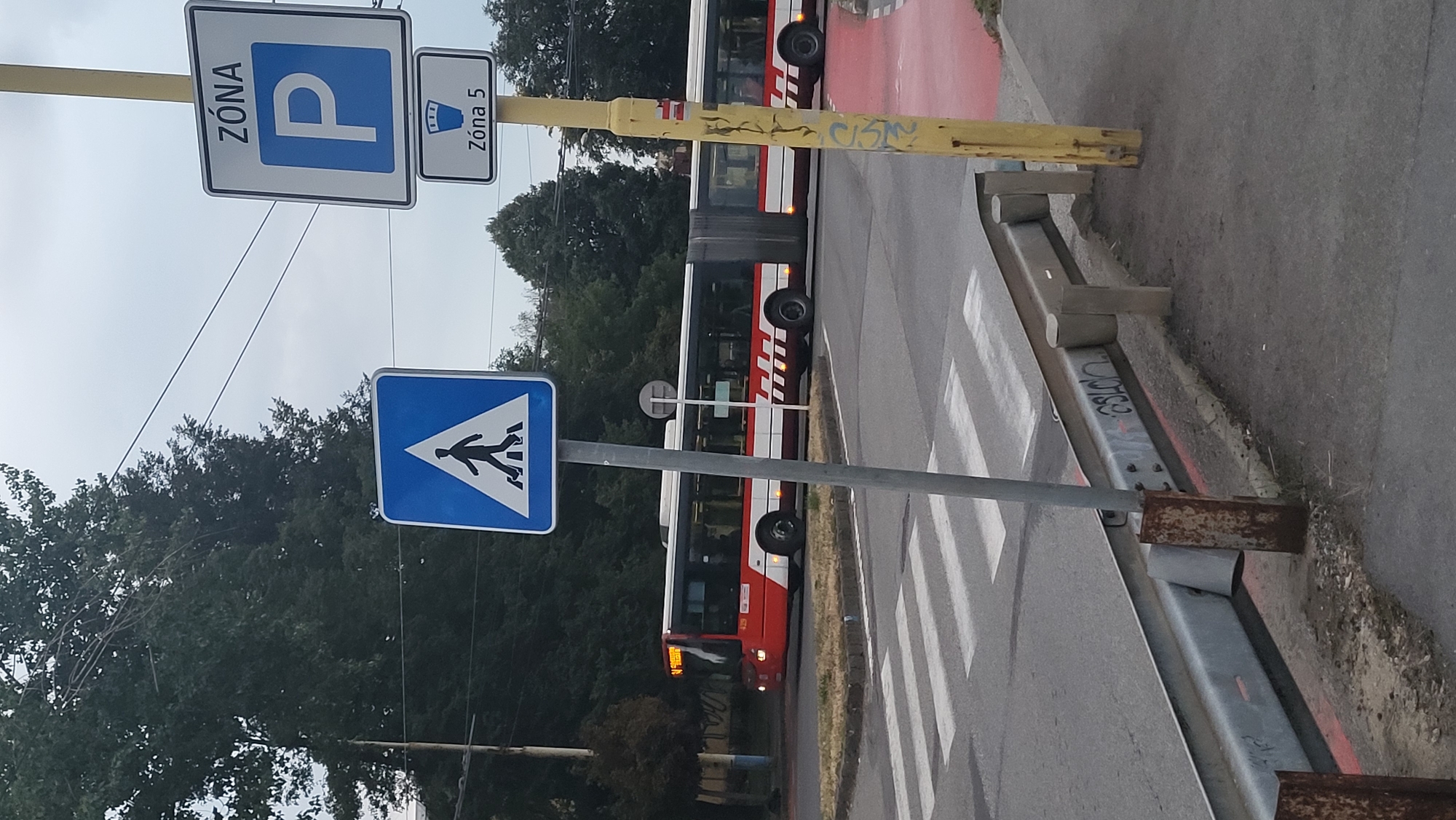 A red and white articulated bus is proceeding through a pedestrian crossing in Prešov, Slovakia. The bus is on the left side of the image, and a pedestrian crossing is in the foreground. There are two traffic signs on poles. One sign, on the left, is a blue square with a white pedestrian symbol. Another sign, on the right, consists of two parts: the top part is a white square with a blue border and a blue letter "P" inside, and the text "ZÓNA" above it. The bottom part is a smaller white rectangle with a blue border, containing a drawing of a parking meter and the text "Zóna 5" below it. A metal guardrail runs along the right side of the image. The sky is overcast.