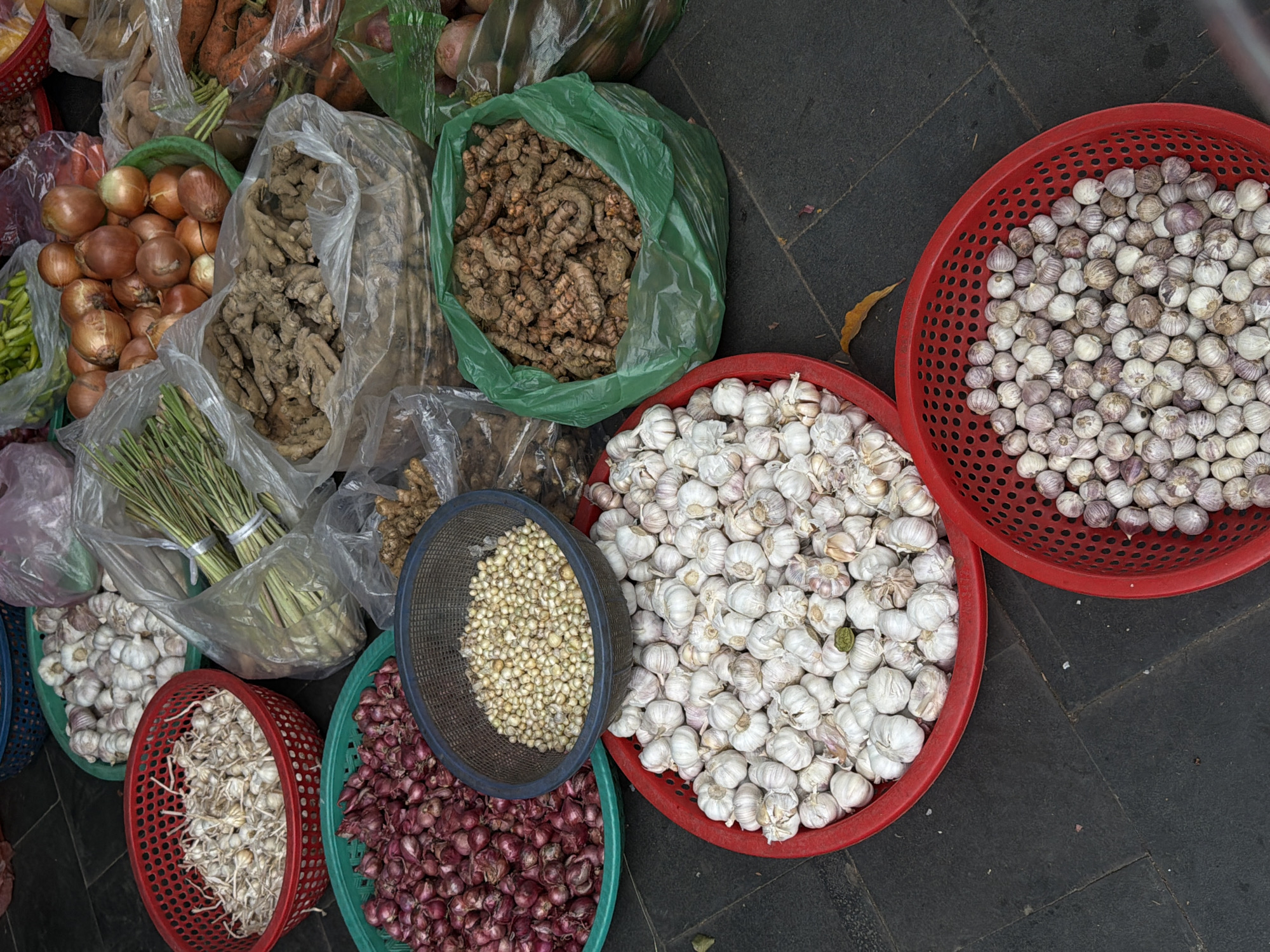 This high-angle, overhead shot captures a rich display of fresh produce, likely at an outdoor market in Hội An Ward, Vietnam. The items are arranged on a dark grey paved surface, presented in a mix of clear plastic bags and colorful perforated plastic baskets.
No people are visible in the frame, focusing entirely on the raw ingredients. The scene is dominated by various spices and alliums. Several types and sizes of garlic are prominent: a large red basket in the upper right is filled with small, purplish-white garlic bulbs, while another large red basket below it holds a heap of larger, white garlic bulbs. A separate dark blue/black basket contains a pile of tiny, pale yellow-white cloves or small bulbs, possibly a different variety of garlic or small onions. A teal-green basket is densely packed with small, deep red shallots or red onions.
Beyond the alliums, other fresh produce includes a large green plastic bag overflowing with gnarled, light brown roots, which appear to be turmeric. Another large clear plastic bag nearby holds numerous pieces of ginger root. Carrots with their green tops are visible in a clear plastic bag in the upper left, alongside larger brown onions. Bundles of fresh lemongrass, tied together, are also seen in clear plastic bags towards the lower left, adjacent to some green chilies.
The arrangement suggests a vendor's stall, showcasing a vibrant array of staple ingredients. The freshness of the produce is evident, with some items still bearing their green stalks. A single dry, brown leaf is visible on the dark paved ground, adding a touch of natural detail. There is no visible text in the image.
