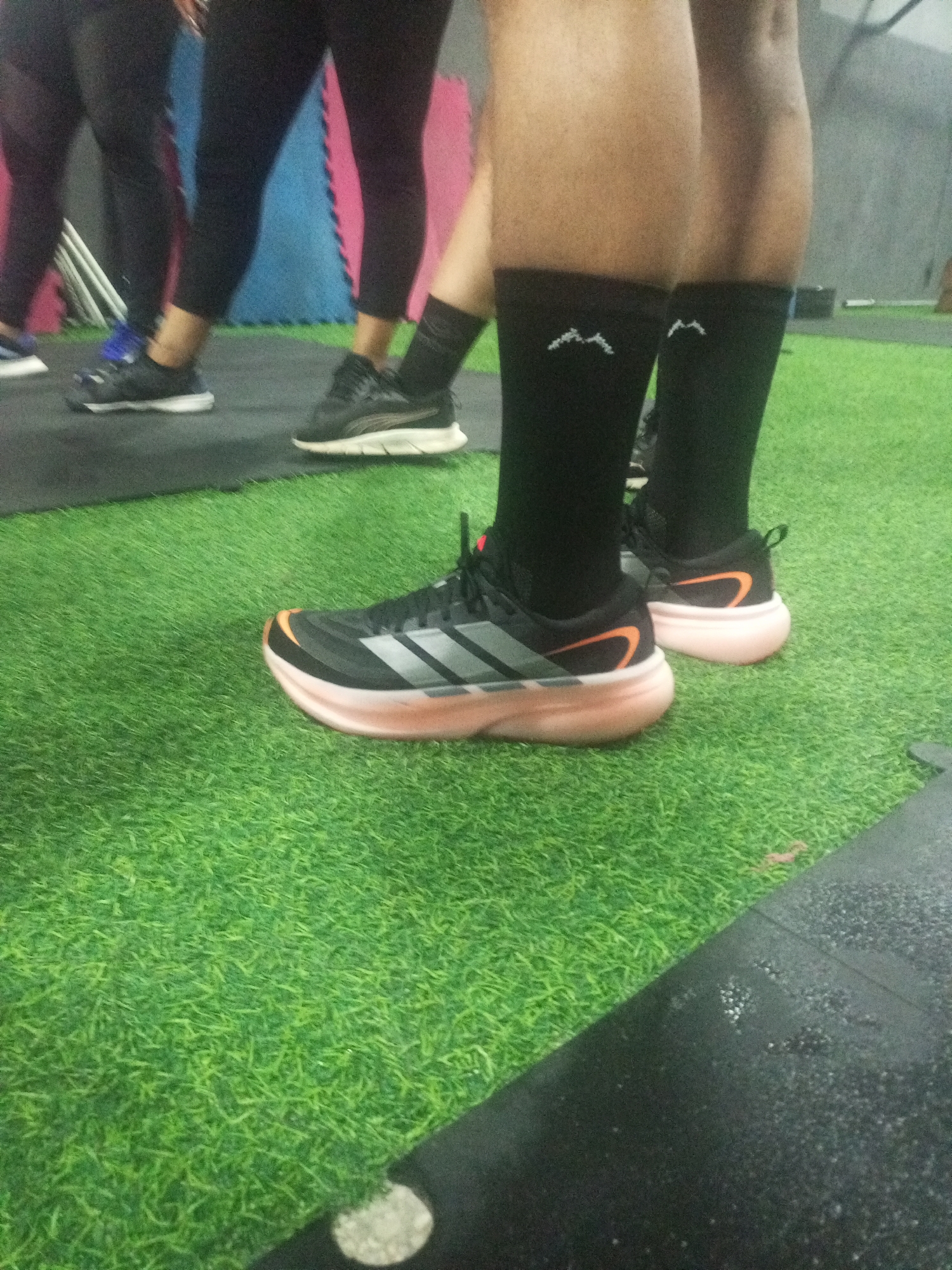 In an indoor gym with artificial turf flooring, a person is standing wearing black athletic socks and black and orange running shoes. The shoes have thick, light-colored soles and grey stripe detailing on the sides. The background shows a wall with pink and blue padding. The image is a close-up of the person's lower legs and feet.