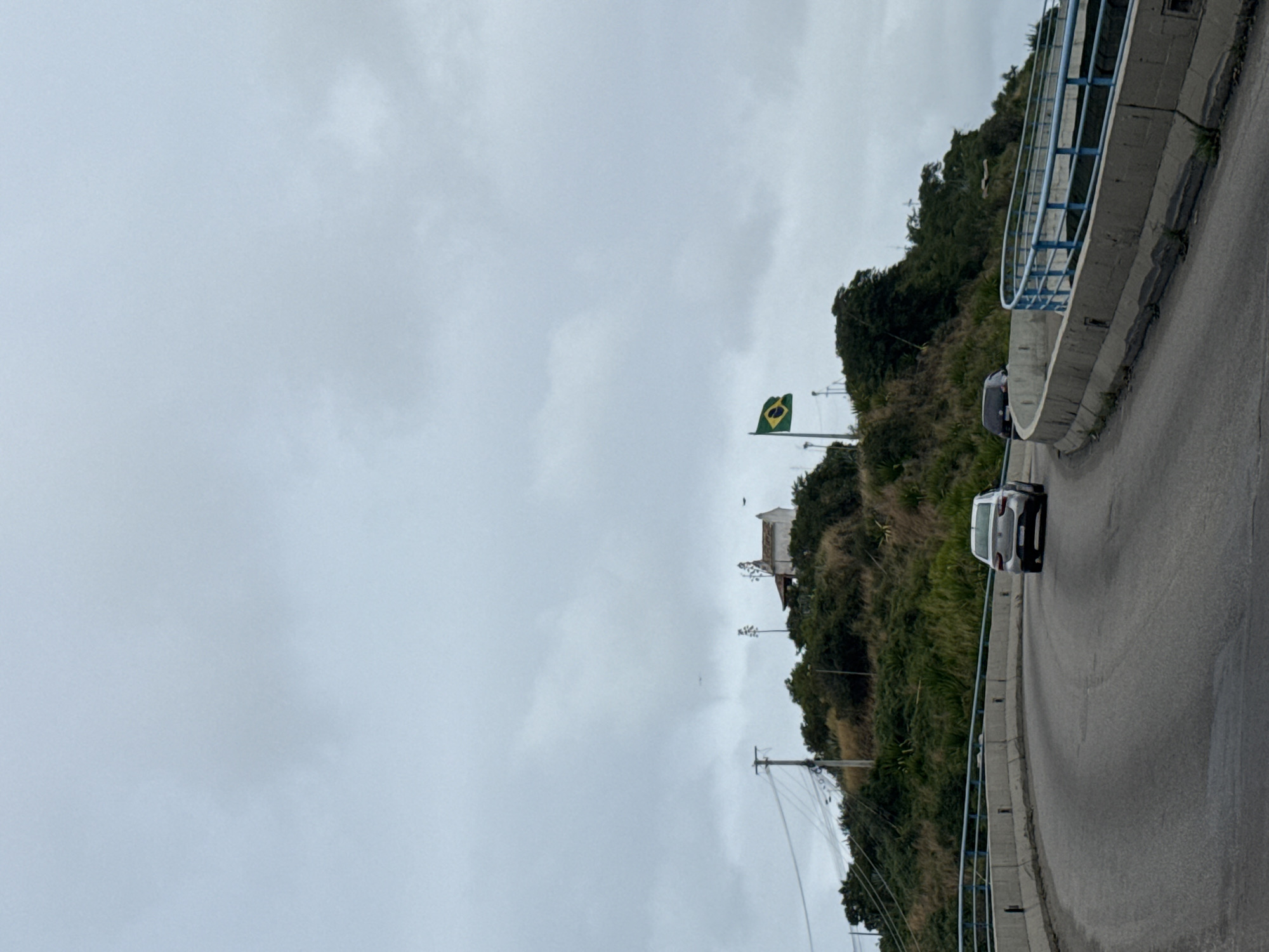 The image shows a paved road curving to the right, with concrete barriers and blue railings lining the edge. Two cars are visible on the road. In the background, a lush green hill rises, topped with a small white building and a Brazilian flag. The sky is overcast and cloudy. The location appears to be a scenic overlook or a road leading to a viewpoint in Cabo Frio, Brazil, likely offering a panoramic view of the city or coastline.