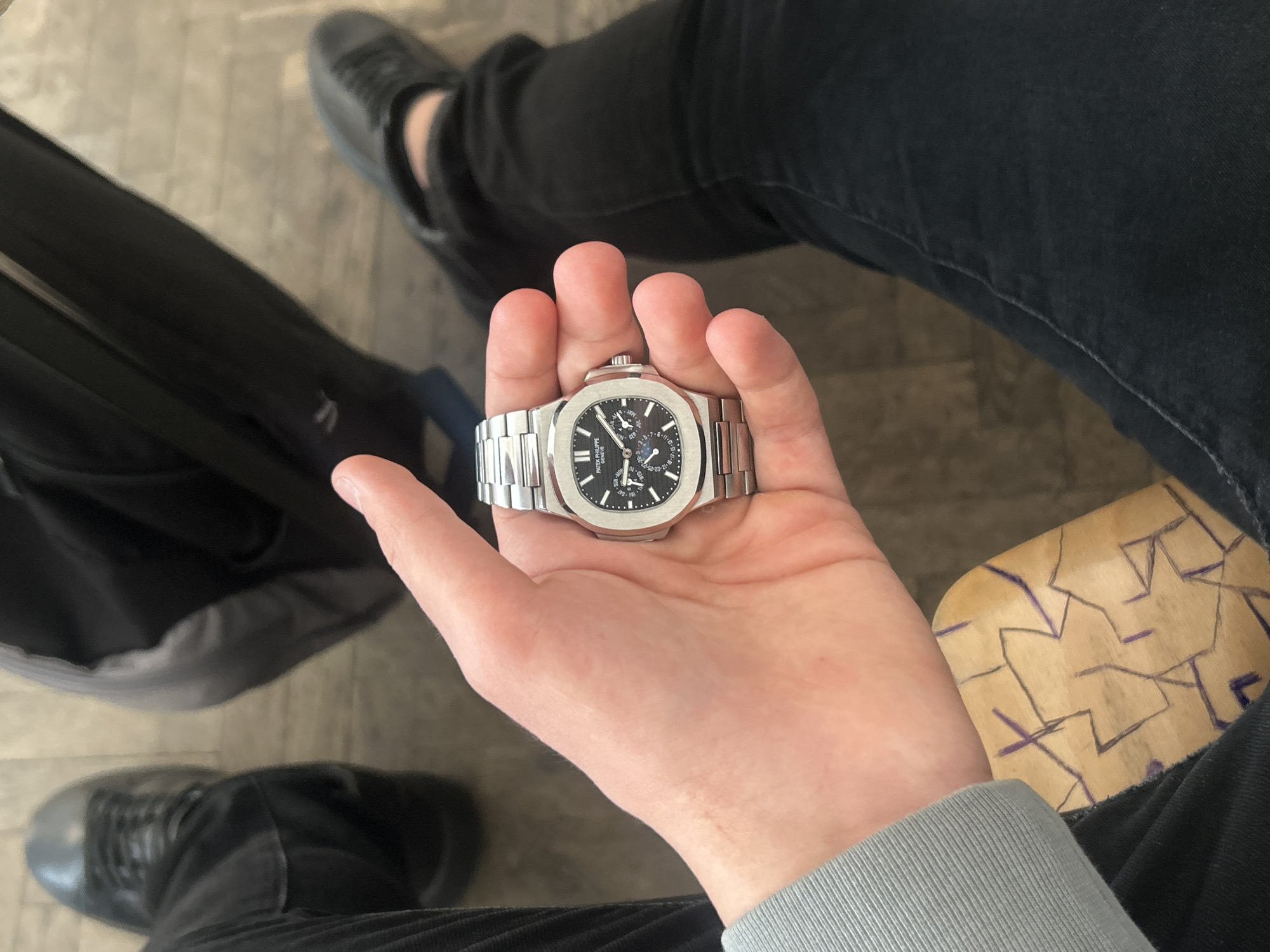 The image provides a close-up, top-down view of a person seated indoors in Zlatograd, Bulgaria. The central focus is the person's right hand, holding a high-end stainless steel Patek Philippe Nautilus watch.
The watch prominently displays a black textured dial with luminous hour markers and hands. It features several complications, including sub-dials for the month (e.g., "MAR", "MAY"), day of the week (e.g., "SAT"), and date (marked with numbers like 3-5-7), along with a moon phase indicator. The brand name "PATEK PHILIPPE GENEVE" is clearly visible on the upper part of the dial. The watch is presented with its integrated steel bracelet.
The individual is wearing black pants and black lace-up shoes, visible in the background along with their left leg. A grey long-sleeved cuff is visible on the person's right wrist. The floor appears to be a light-colored, possibly tiled or concrete surface. To the right of the hand, a portion of a wooden surface with abstract purple markings, resembling doodles, suggests a chair or table the person is seated on or near. A dark bag or backpack strap is partially visible on the left side of the frame. The lighting suggests a well-lit indoor environment. The activity captured is the presentation or photography of the luxury watch.