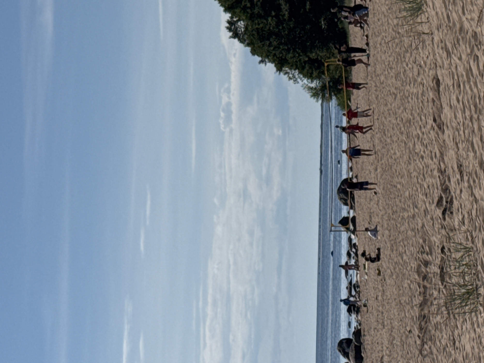 The image depicts a beach scene in Zelenaya Roshcha, Russia. The foreground is a sandy beach with sparse vegetation. In the midground, a group of people are playing volleyball. There are several other individuals scattered across the beach, some sitting and others standing. The background features the sea, a rocky shoreline, and a dense green forest. The sky is blue with scattered clouds. The scene appears to be a leisure activity on a sunny day.