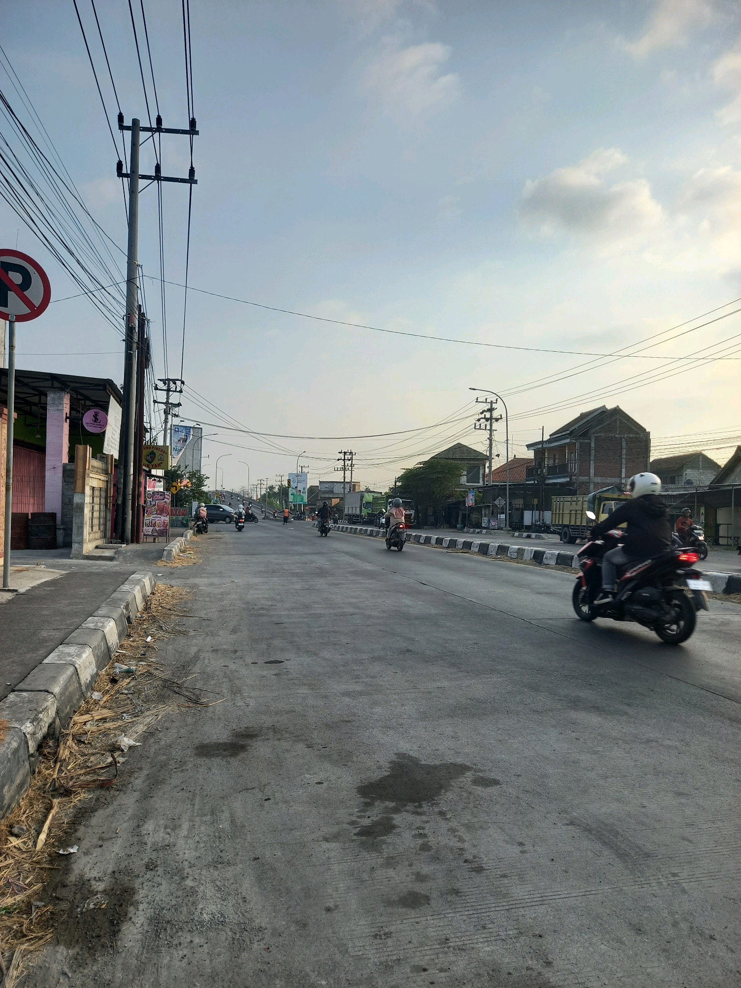 The image depicts a street scene in Bedah Lawak, Indonesia, during what appears to be late afternoon or early morning due to the soft lighting and long shadows. The road is paved and shows signs of wear, with some patches and debris along the curb. Several motorcycles are visible, with one rider in the foreground on a black and red motorcycle, wearing a white helmet and a dark jacket. Other motorcycles are further down the road, some with riders, suggesting moderate traffic.
On the left side of the street, there is a sidewalk with a curb and some dry vegetation and litter. Adjacent to the sidewalk are low-rise buildings, one with a pink and grey facade and an awning. A "no parking" sign with a red circle and a diagonal bar over a "P" is visible on a pole. Overhead, a dense network of power lines and utility poles crisscrosses the sky, characteristic of urban infrastructure in Indonesia.
Further down the road, there are more buildings, including some houses with red tile roofs and brick structures. A large truck with a yellow container and a green truck are also present on the right side of the road. The sky is a pale blue with some scattered clouds. The overall impression is a typical street in a populated area of Indonesia, with a blend of commercial and residential buildings, active transportation, and abundant electrical infrastructure.
