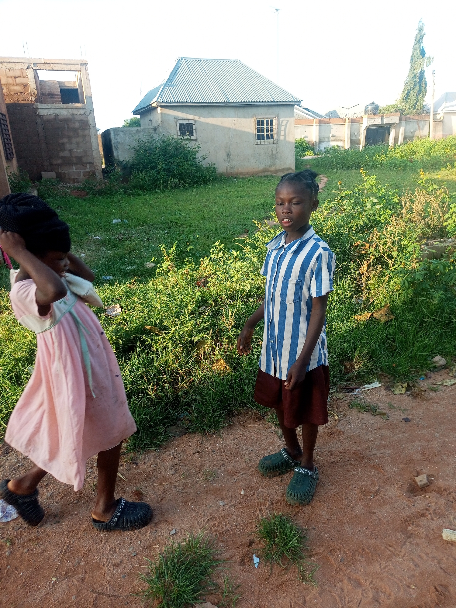 In an outdoor scene, two young girls stand on a dirt path in Tempure, Nigeria. The girl on the left, wearing a light pink dress and black head covering, is walking away from the camera, her hands near her head as if adjusting her head covering. The girl on the right, facing the camera, wears a blue and white striped short-sleeved shirt and a dark red skirt. Both girls are barefooted in the foreground. Behind them, a low-lying building with a corrugated metal roof is visible, along with other structures in the background. Green grass and vegetation are scattered throughout the scene. The lighting suggests it is daytime, possibly late afternoon, with a warm glow.