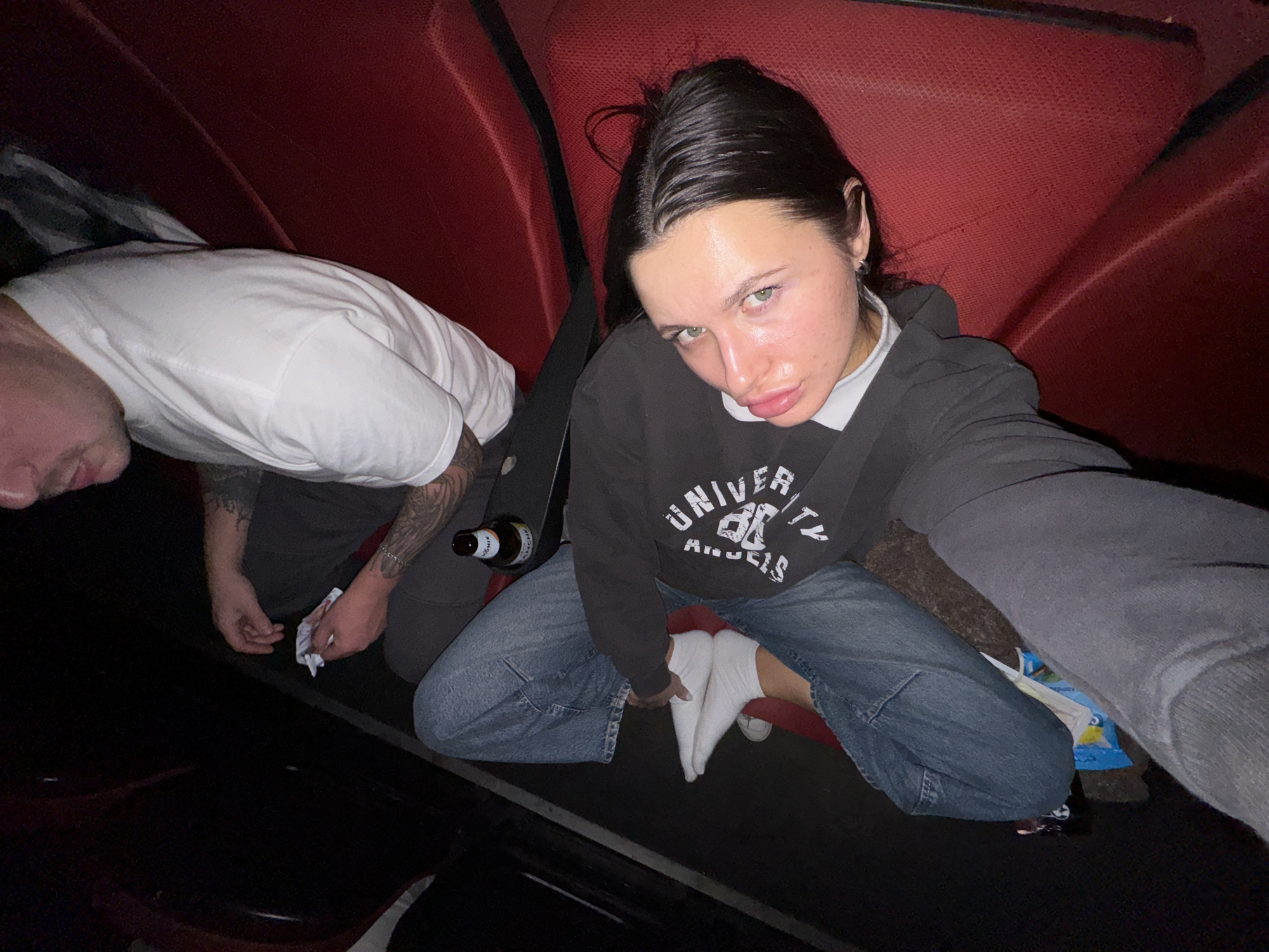 The scene depicts two individuals in a dimly lit interior, characterized by red upholstered seating, possibly a vehicle or private booth. No identifiable features confirm the specific location as Lesnoi Gorodok, Russia.
One subject, a male, is partially visible from the chest upwards, seated on the red upholstery and facing away from the camera, slightly turned to his left. He wears a white short-sleeved t-shirt and dark pants. Tattoos are present on both forearms. His right hand holds a crumpled white item. A brown glass beer bottle, partially labeled "OHIX," rests near his left leg on an adjacent surface.
The second subject, a female, is prominently featured, reclining on the red upholstery and facing directly towards the camera. She has dark hair, light-colored eyes, and full lips. She is dressed in a dark hooded sweatshirt with white lettering (partially visible as "UNIVER"), a white t-shirt underneath, blue jeans, and white socks. Her left arm is extended towards the camera, indicating she is operating the device capturing the image. Her legs are crossed at the ankles, resting on a dark surface. A small blue and white package is visible beneath her legs. The individuals are in close proximity within the confined space.