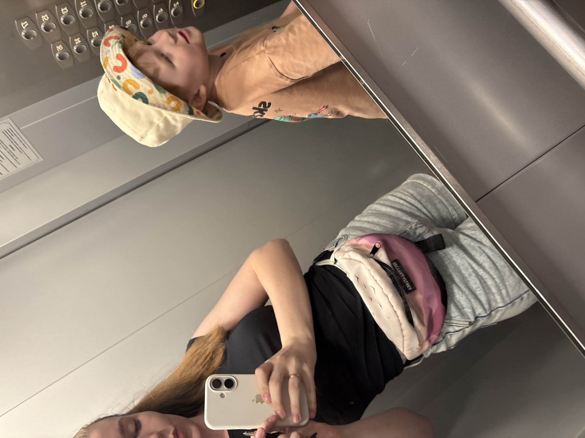 The image is a selfie taken in an elevator. A woman is holding a white iPhone, taking the photo. The reflection in the elevator door shows a child in a tan shirt and colorful hat near the elevator buttons. The woman is wearing a black shirt and light wash jeans with a pink and white fanny pack. The location is unknown and not mentioned in the image.