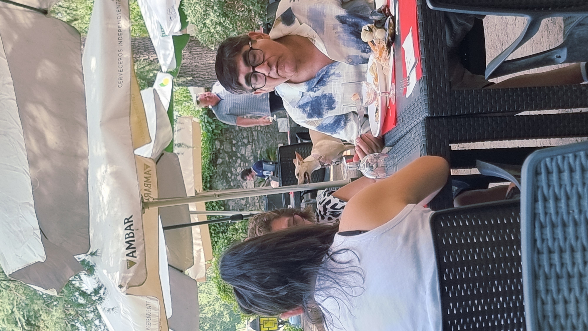 The image depicts an outdoor dining scene at a restaurant, likely located in or near Besalú, Spain, suggested by the presence of umbrellas with "AMBAR" branding, a Spanish beer. In the foreground, three individuals are seated at a black, woven table. The person on the far right, a man wearing glasses and a blue and white patterned shirt, is in the process of eating, with a fork raised to his mouth and food on his plate. In front of him are a white plate with food, a wine glass, and cutlery. To his left, a woman with dark hair, wearing a white tank top, is partially visible, her arm resting on the table. Between the man and the woman with dark hair, another woman is seated, wearing a patterned top and glasses, also looking towards the table. A light-colored dog with a pink collar stands near the table, looking towards the food. In the background, several other umbrellas are visible, along with trees and stone walls. Two other men are further in the background; one is wearing a blue striped shirt and appears to be walking, while another is seated at a table with a laptop. The overall setting suggests a relaxed, sunny day at an outdoor restaurant or cafe.