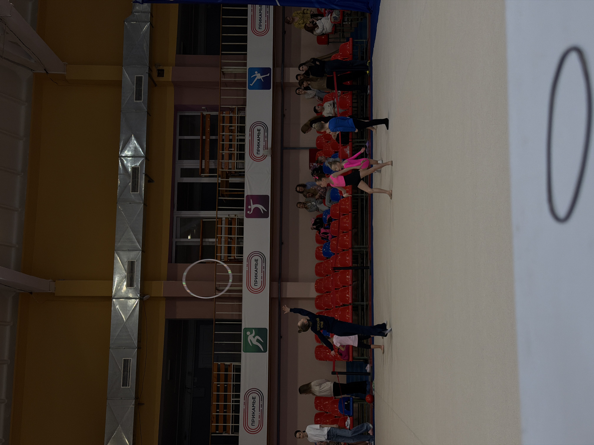 The image shows a gymnastics competition taking place indoors in Perm, Russia. Two young gymnasts are performing on a white mat. One gymnast is wearing a dark blue leotard, and the other is in a pink and black leotard. A hoop is in the air near the edge of the mat. Spectators are seated in red chairs along the side of the mat, watching the performance. Banners with Cyrillic script are behind the seating area. The room has yellow and pink walls with ductwork running along the ceiling.