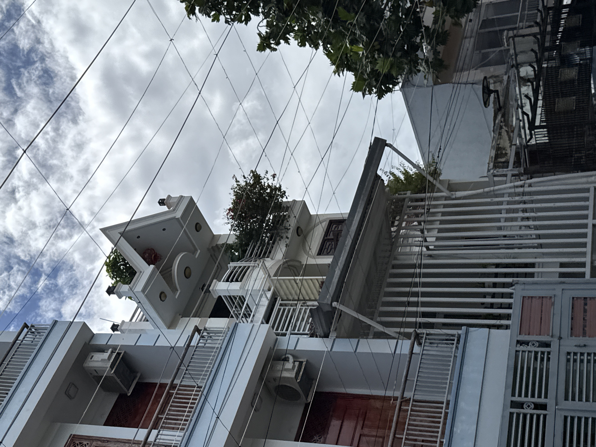 The image shows a close-up, low-angle view of a multi-story building exterior in Nha Trang, Vietnam, under a cloudy sky. The building is light blue with white trim and features several balconies with metal railings. One balcony on the left has a red door. An air conditioning unit is visible on the side of the building. Above this level, another section of the building has a white, gabled roof-like structure with decorative elements and potted plants. The image is dominated by a complex network of dark electrical wires crisscrossing the sky. To the right, the branches of a leafy green tree are visible. In the foreground, a metal gate with horizontal slats and some red accents is partially visible at the bottom. The scene suggests an urban residential or commercial area.