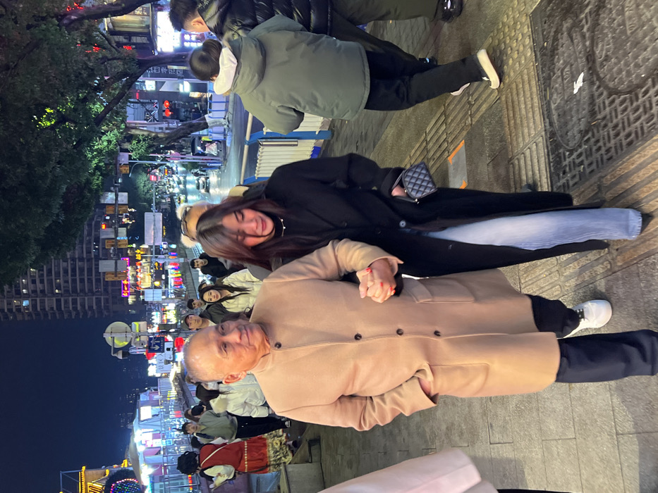 This image depicts a nighttime urban street scene in Yuzhong District, China. In the foreground, an older Asian male and an Asian female walk side-by-side on a paved sidewalk. The male, with a bald head and light skin, wears a knee-length tan overcoat with a mandarin collar, four dark buttons, and two front pockets. He has his left hand in his coat pocket and holds the female's right hand with his right hand. He wears dark trousers and white sneakers.
The female has long dark hair and wears a long black overcoat, light blue wide-leg jeans, and dark pointed-toe shoes. Her left hand holds a small black quilted handbag with a gold-colored chain strap. Her face displays visible moisture around her eyes and a distressed expression. She wears a thin silver-colored necklace.
They are positioned on a grey paved sidewalk that features metal utility covers. To their right, a low white and blue metal barrier separates the sidewalk from a multi-lane street. The street shows blurred vehicle headlights and taillights, indicating active traffic. The mid-ground and background are populated by numerous pedestrians, predominantly dressed in winter clothing. One individual on the far left wears a red vest over a white garment and a patterned skirt.
The environment is densely urban, characterized by multiple multi-story buildings and high-rises lining the street. These structures are extensively illuminated with various colorful neon and LED signs and decorative lighting, creating a bright urban nightscape. Streetlights, traffic signals, and green-leafed trees are also visible along the street.