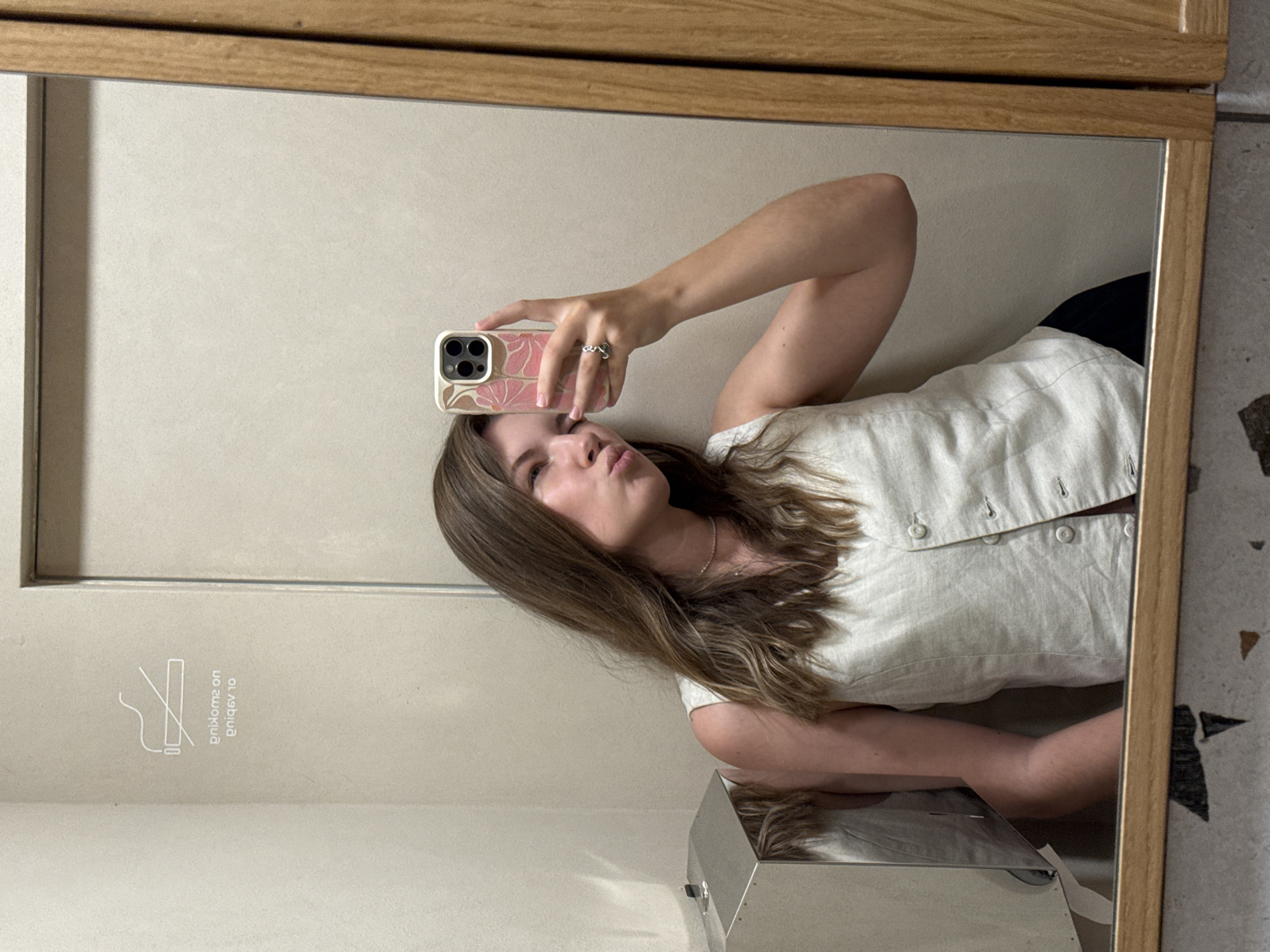 A young woman with long, light brown hair is taking a mirror selfie in what appears to be a public restroom in Vracar Urban Municipality, Serbia. She is wearing a light-colored, sleeveless button-up vest over dark pants and has a ring on her right ring finger. The woman is leaning against a wall and holding a smartphone with a floral-patterned case. Her left arm is bent, and her hand is resting on her cheek. To the left of the frame, a stainless steel trash can is visible, and a "No Smoking" sign is affixed to the wall above it. The background is a neutral-toned wall, and the mirror reflects the woman and the surrounding environment. The floor has a speckled pattern. The lighting is ambient, suggesting indoor lighting.