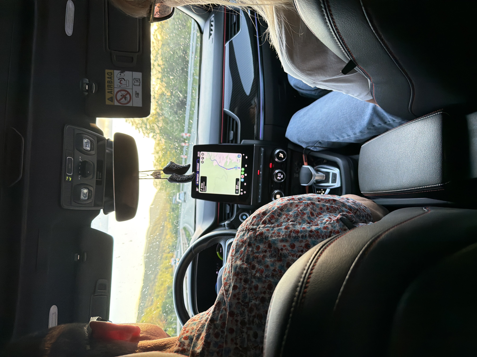 A view from the back seat of a car, possibly in motion. A person is seated in the driver's seat. A GPS screen is visible, displaying a map that suggests the location is near Novi han, Bulgaria. A second person, wearing blue jeans and a light-colored shirt, is in the passenger seat. The car's interior is black with red stitching on the seats.
