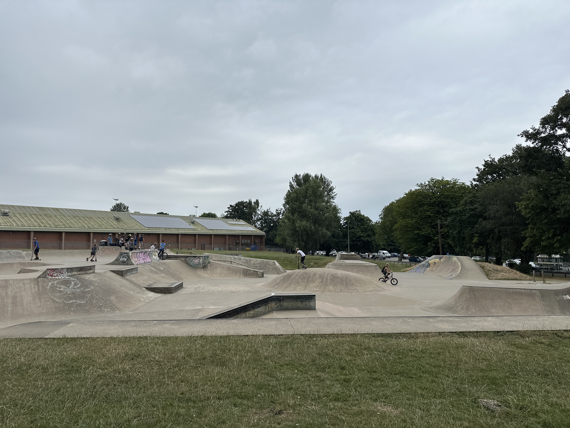 The image depicts a skatepark with several individuals engaged in activities. The foreground is dominated by a grassy area. The middle ground features a complex concrete skatepark with various ramps, bowls, and obstacles. Several people are present in the skatepark. On the far left, a person on a scooter is moving across the concrete. Near the center, a person on a scooter is on a mound, and to their right, another person on a bicycle is riding on a ramp. In the background, a building with a green roof and solar panels is visible. Trees and parked cars are also present in the background, suggesting an outdoor urban or suburban setting. The sky is overcast. The image is likely taken in Wakefield, United Kingdom, given the mention of the city.
