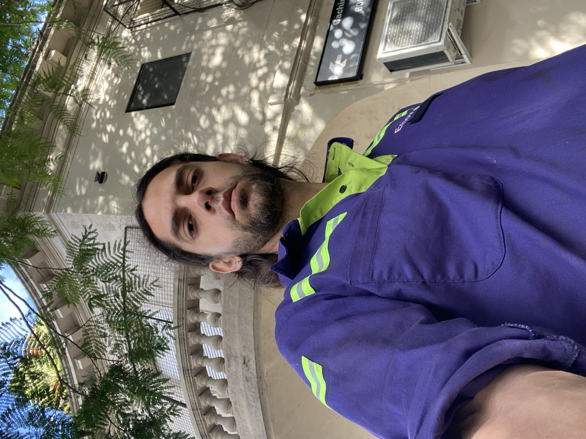 The image is a selfie taken by a man from a low, upward-looking angle, giving a unique perspective on an urban setting.
The main subject is a man with long dark hair, a dark beard, and mustache, looking directly at the camera with a neutral expression. He wears a deep purple or indigo work shirt or jacket featuring a bright lime green collar and prominent lime green reflective strips on the shoulders and arms. A small silver earring is visible in his left ear.
In the background, a light-colored building façade, possibly beige or white, stands tall. It features an ornate balcony railing with classic balusters, a dark rectangular window, and a white air conditioner unit mounted on the wall. Lush green foliage from nearby trees partially frames the scene, casting intricate dappled shadows across the building's surface. Through the leaves, patches of clear blue sky are visible, indicating bright, sunny daytime weather.
On a black sign attached to the building, partially obscured, the text "Gachina" and "600" are discernible, likely indicating a business name and a street number. The architecture and urban elements are consistent with a city environment like Buenos Aires, Argentina. The overall impression is a casual, sunlit moment captured in an older urban streetscape.