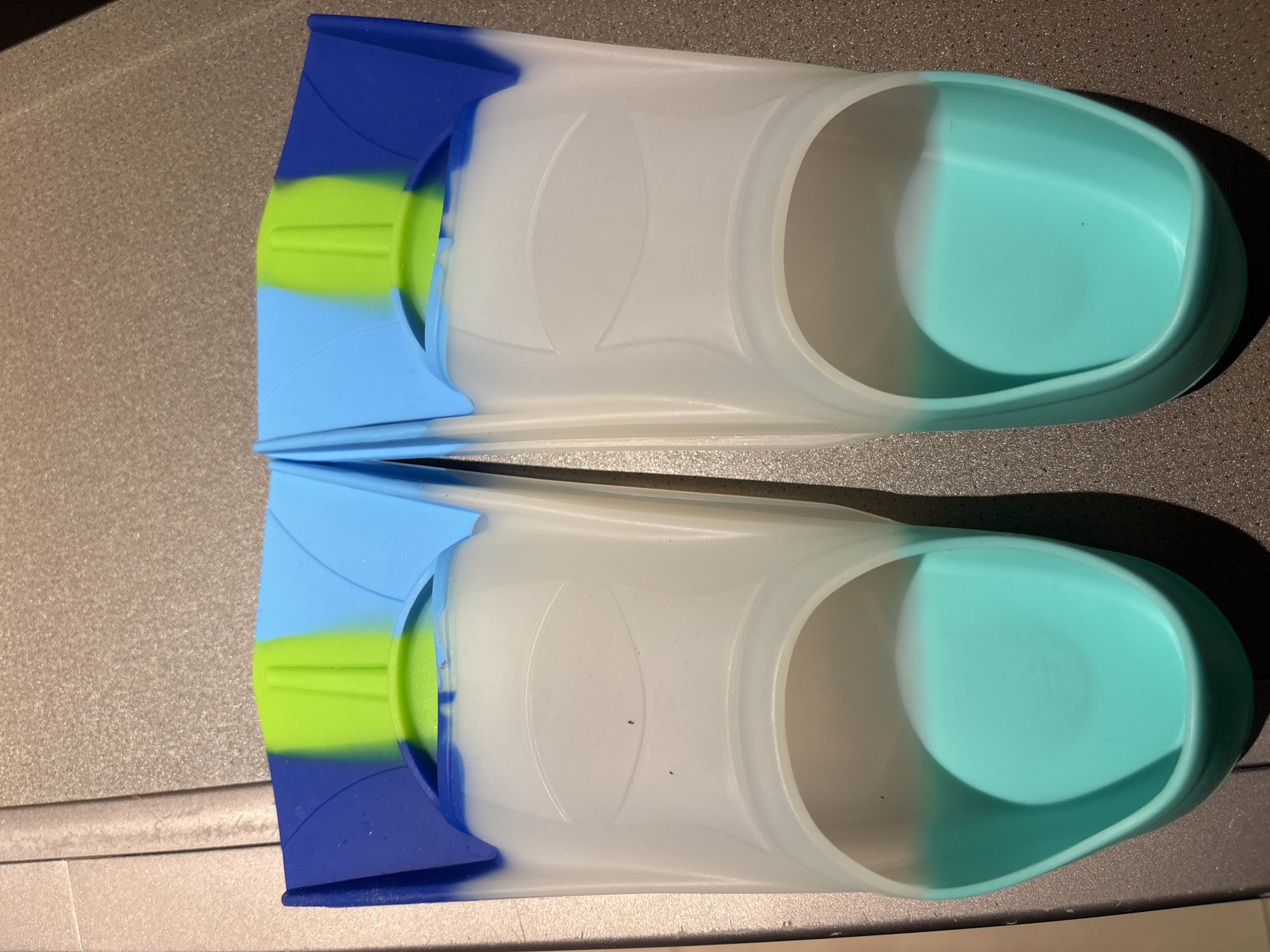 A pair of snorkeling fins are placed on a textured, speckled gray surface, possibly a countertop. The fins are made of a semi-transparent white material with a teal-colored foot pocket and a blue and lime green colored blade. They are positioned side-by-side, facing upwards, with the blades slightly angled away from each other. The background shows a thin, lighter colored vertical strip on the left, suggesting a wall or edge. The lighting casts subtle shadows from the fins onto the surface. This image does not contain any information to suggest a location in Dubai, United Arab Emirates.