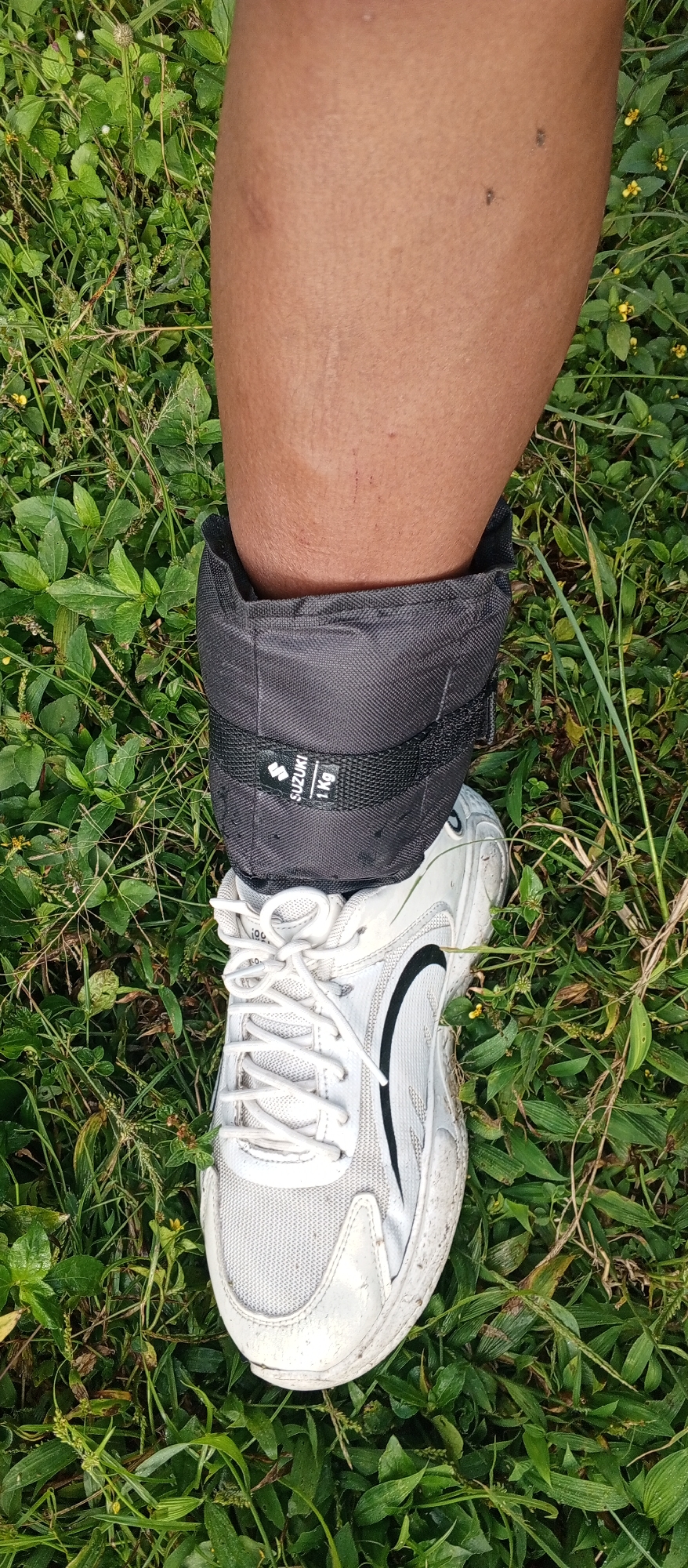 A person's lower right leg and foot are positioned vertically on a verdant, overgrown patch of grass and broadleaf plants in Weddagala, Sri Lanka. The leg displays bare, medium-brown skin with visible pores, a light tan line around the ankle, and several small blemishes or insect bites on the calf. A black fabric ankle weight, marked with a white "SUZUKI 1 Kg" label, is secured around the ankle. Below the ankle weight, a white athletic shoe with black accents and white laces is worn. The shoe's surface appears slightly worn and soiled, consistent with outdoor use. The surrounding ground is densely covered with various green foliage, including slender blades of grass and wider leaves, with some small yellow flowers interspersed throughout.