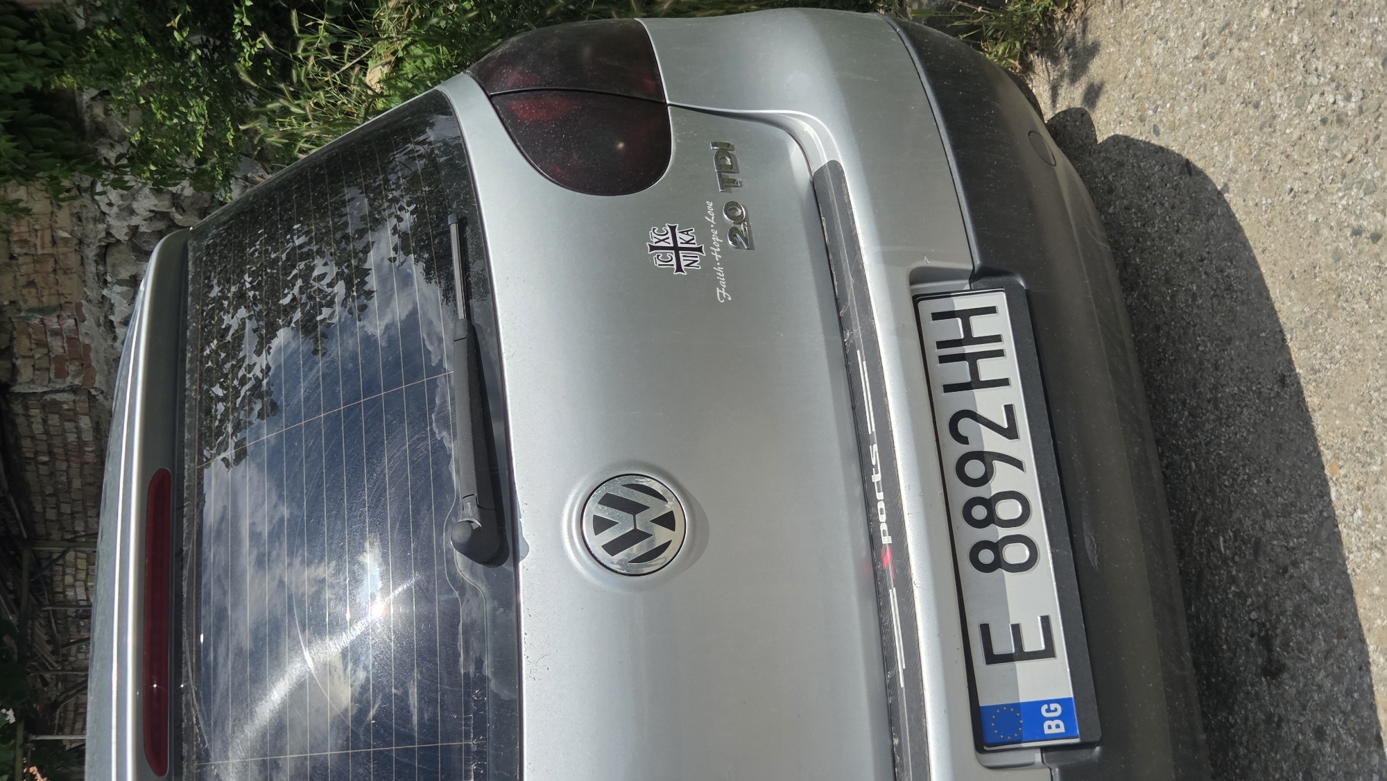 The image shows the rear of a silver Volkswagen hatchback parked outdoors. The car is facing the camera. A license plate with the characters "E 8892 HH" is visible, along with the European Union and Bulgarian flags on the left side of the plate. A sticker with a cross and the letters "IC XC NIKA" is on the right side of the car, above another sticker that reads "Faith Hope Love 2.0 TDI". The car is parked on a gravelly surface, and there is a brick wall and greenery in the background. The sky is visible through the car's rear window, reflecting clouds and sunlight.