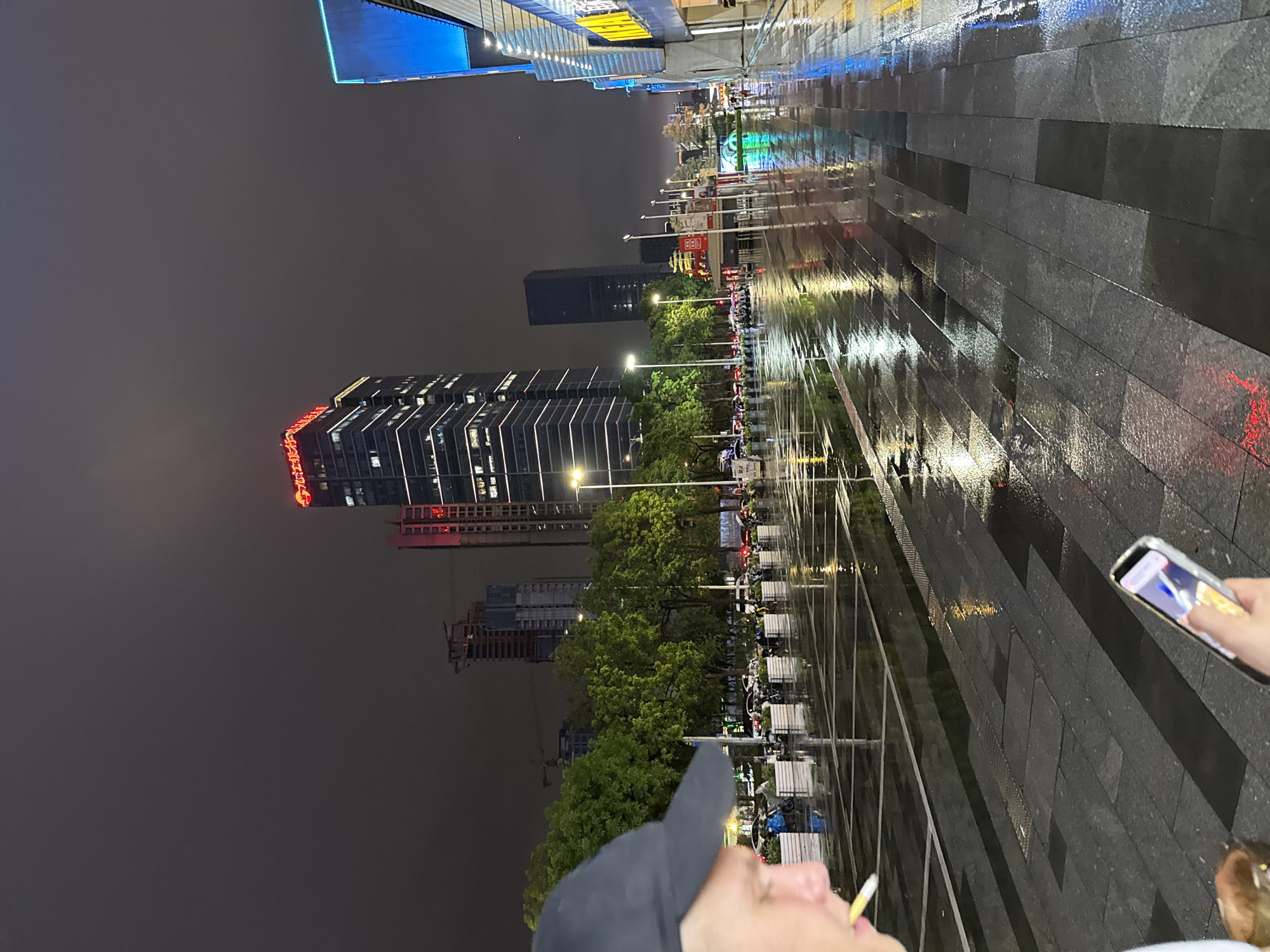 In Tianhe District, China, at night, a person wearing a dark baseball cap is captured in a close-up profile view, looking down at a smartphone held in their right hand. The person appears to be smoking, with a cigarette in their mouth, and a beverage container is visible near their left hand. The scene unfolds on a wet, paved street reflecting the neon lights of surrounding buildings and streetlights. In the background, a cluster of modern skyscrapers, some illuminated with bright lights and signage, dominates the skyline. Trees line the street, and a row of parked motorcycles is visible. The wet pavement and reflections suggest it has recently rained, creating a glossy, urban nighttime atmosphere.