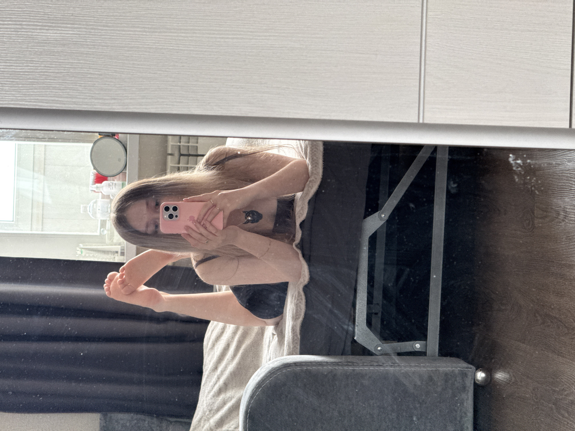 The image depicts a mirror selfie taken in an indoor residential setting. A female individual, identified by her long, light brown hair, is lying prone on a bed, facing towards the viewer's right, and holding a pink smartphone to capture her reflection. Her face is largely obscured by the phone, which displays multiple camera lenses. She is wearing a dark top, possibly a bra or camisole, and dark shorts or underwear. A dark pendant, shaped like an animal head, is visible around her neck. Her legs are bent upwards at the knees, with her bare feet visible against a dark headboard or wall behind the bed. Rings are present on her right hand, which holds the phone, and a thin bracelet is on her left wrist.
The bed features a dark frame and is partially covered by a light-colored, textured blanket or throw. To the left of the individual, a dark curtain hangs. Behind her, a window is visible, through which daylight enters. On the windowsill, a clear plastic bottle, a white pump bottle, and a round mirror are present. Below the window, a white radiator or heating unit is partially visible. To the extreme left of the frame, the armrest and a spherical metallic leg of a dark gray upholstered armchair or sofa with white stitching are visible. The flooring is dark wood or laminate. On the far right side of the image, outside the mirror's reflection, a light-colored, vertically paneled wardrobe or cabinet is present. The specific location within the city of Chaikovsky, Russia, is not ascertainable from the visual information provided.