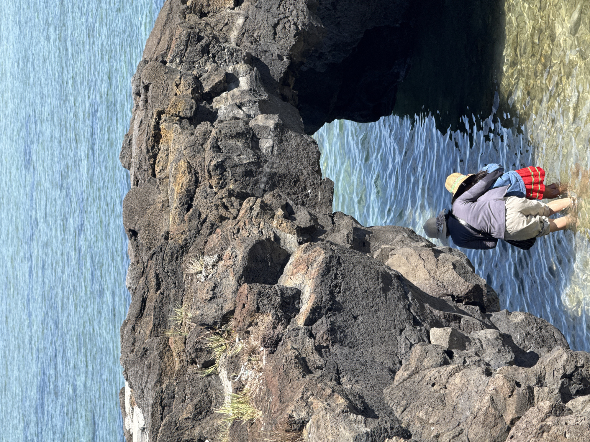 Two people, an adult and a child, stand in shallow, clear water near a rocky archway on the coast of Lý Sơn Island, Vietnam. The adult, wearing a hat and a grey jacket, has their arm around the child, who is wearing a straw hat and a red skirt. The water is calm and reflects the sunlight. The rocky archway is dark and rough, with sparse vegetation growing on top. In the background, the ocean stretches out to the horizon, its surface rippled by a gentle breeze. The scene suggests a peaceful moment of exploration and connection with nature.