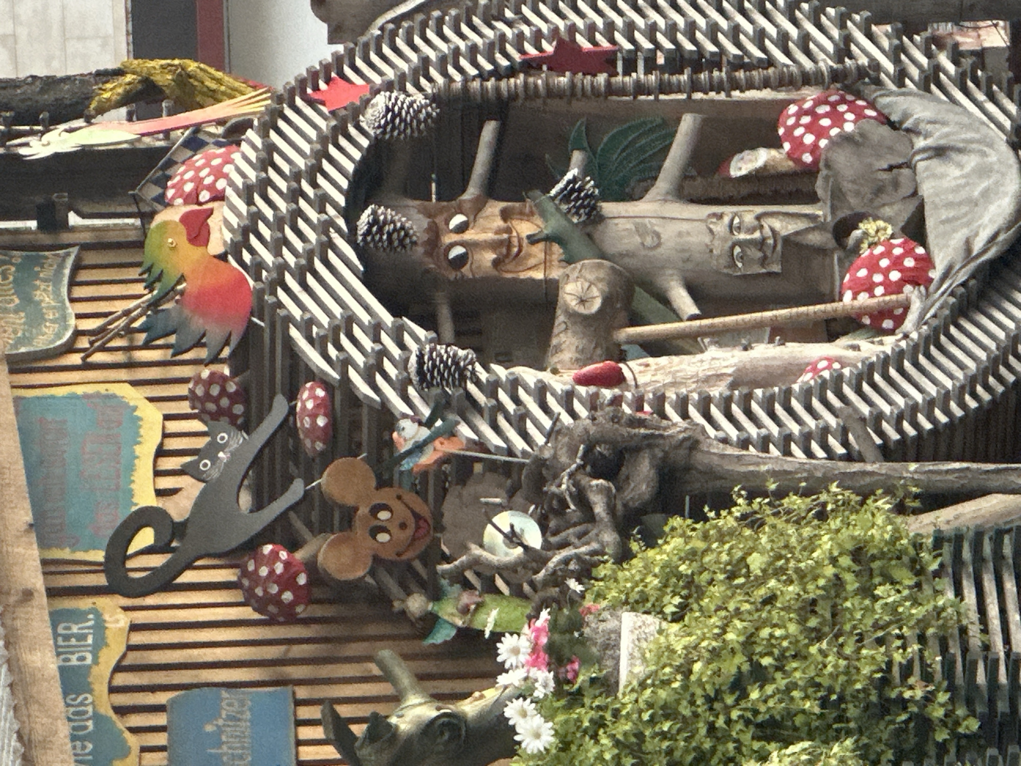 This image depicts a whimsical outdoor display, likely in Günzburg, Germany, featuring a variety of folk art and decorative elements. The scene is dominated by a central, carved wooden figure resembling a tree trunk with a smiling, anthropomorphic face. This figure is framed within an oval structure made of dark wooden slats arranged in a diagonal pattern. Surrounding the central figure are various whimsical objects, including red and white spotted mushroom-shaped decorations, pinecones, and a small, carved wooden bird with colorful plumage.
To the left of the central display, a black metal cutout of a cat with large, round eyes is prominently featured, its tail curled upwards. Below the cat, a wooden cutout of Mickey Mouse's head with a wide smile and a protruding tongue is visible. Further to the left, a statue of a goat or similar animal head is partially obscured by lush green foliage and white and pink flowers in a planter. A small, green fairy-like figurine holding a pink object can also be seen.
A sign with blue text on a yellow background reads "wie das BIER." Another blue sign with white text says "schnitzer." The background consists of vertical wooden planks. The overall impression is one of a playfully decorated exterior, possibly a shop or a garden feature, showcasing handcrafted artistic elements.