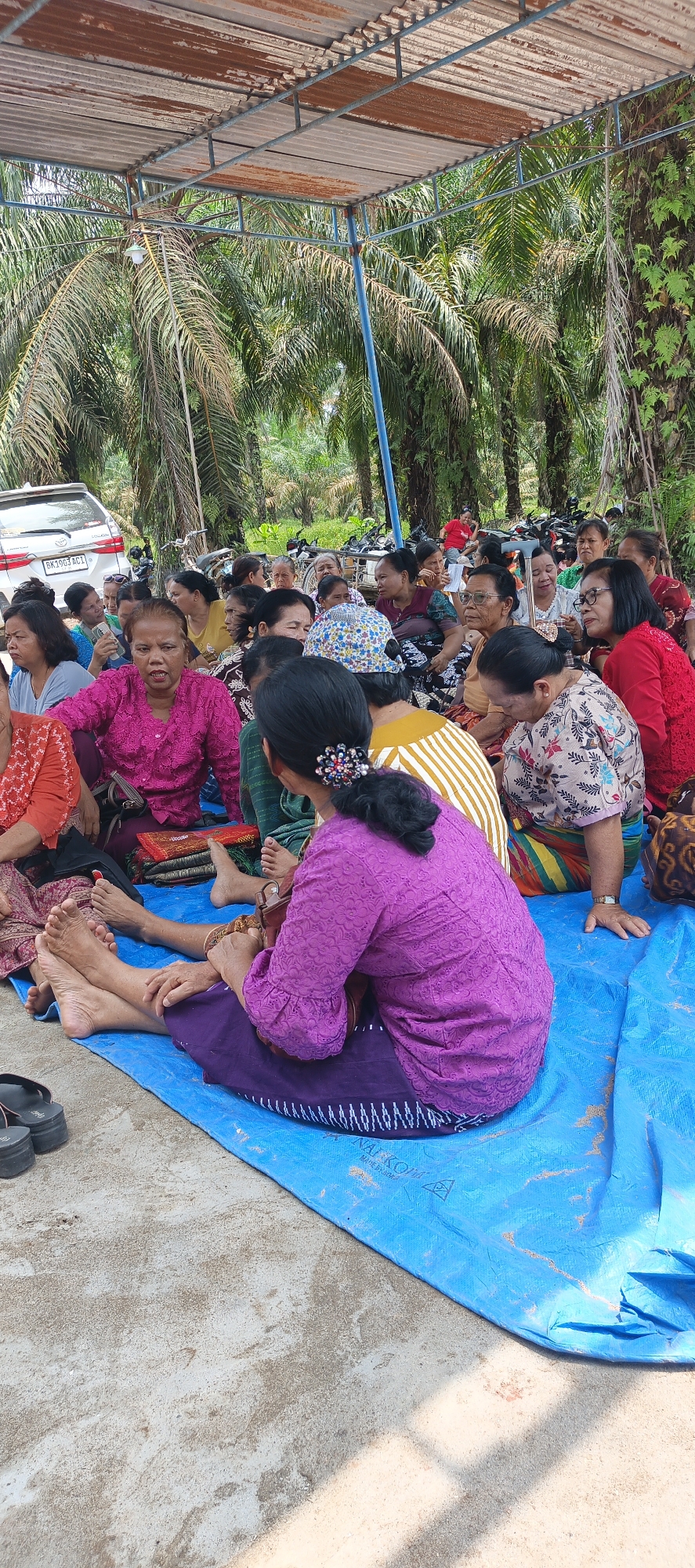 The image depicts a gathering of approximately 30 people, predominantly women, seated on a blue tarp outdoors. The setting appears to be a rural or semi-rural area, with palm trees and other foliage visible in the background. A rusty, corrugated metal roof supported by blue metal poles provides partial overhead cover. In the background, several motorcycles are parked, and a white car with license plate BK 1903 ACI is visible. The people are dressed in various colorful outfits, with some wearing traditional Indonesian attire. Some individuals are looking towards the camera, while others are engaged in conversation. The overall atmosphere suggests a community event or meeting. The presence of the palm trees and the vehicle license plate strongly suggest the location is in Indonesia, and specifically Negeri Lama.