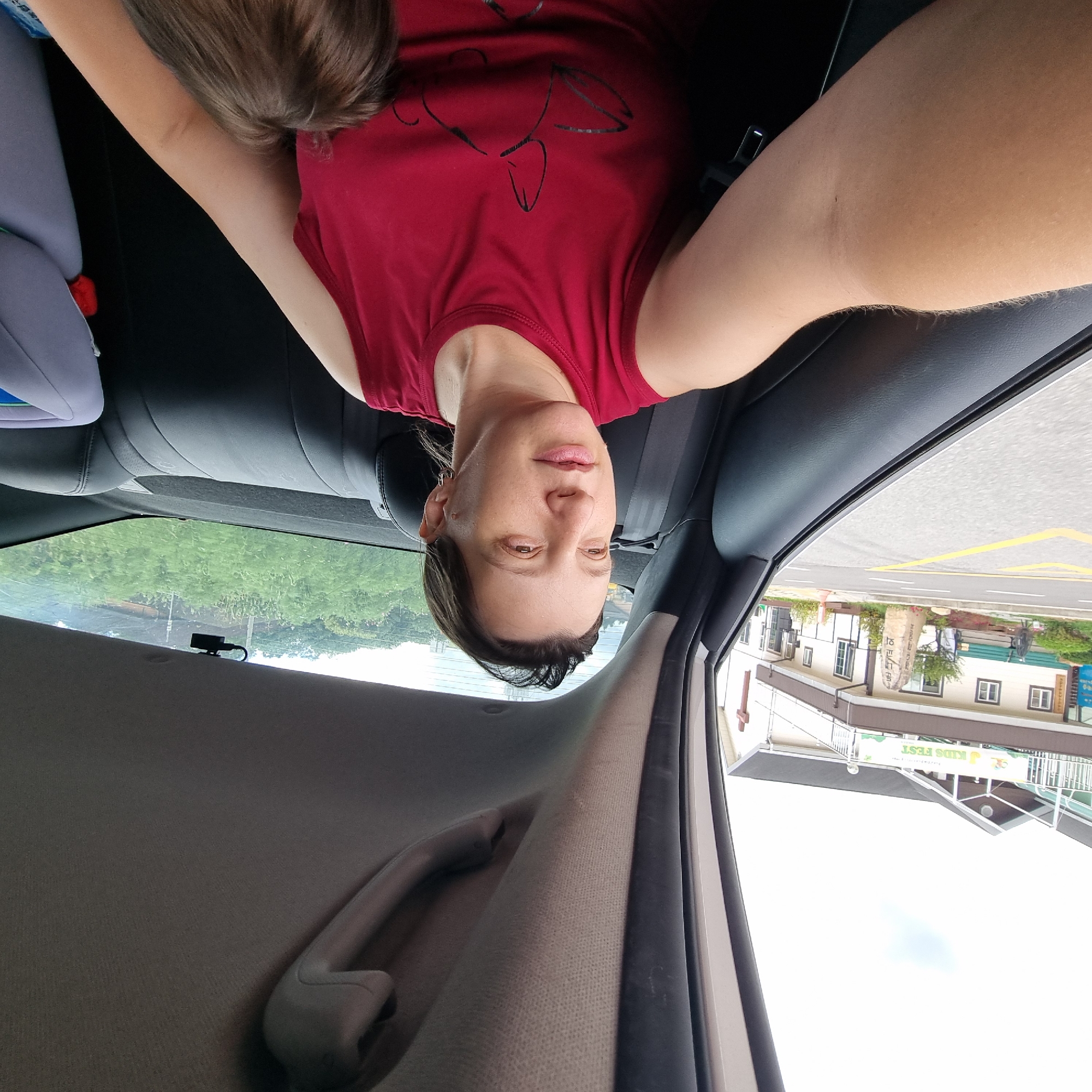 A medium shot captures a woman with medium-length brown hair and fair skin, wearing a sleeveless red top with a black graphic print, seated in the back of a car. She is looking to her left, with her mouth slightly open and her eyes focused ahead. Her left arm is extended, and her right arm is partially visible, holding a small child whose brown hair is peeking out from behind her. The car's interior is visible, including the black leather seats and the grey headliner with a grab handle. Through the car window, a building with a "KIDS FEST" banner is visible, along with a road marked with yellow lines, a stone monument with Korean writing, and a white cross on the building, suggesting a church or religious institution. Lush green trees are visible in the background. The image is taken from a slightly high angle, looking down at the woman. The overall scene suggests a moment captured during a car journey in the city of Dunnae-myeon, South Korea.