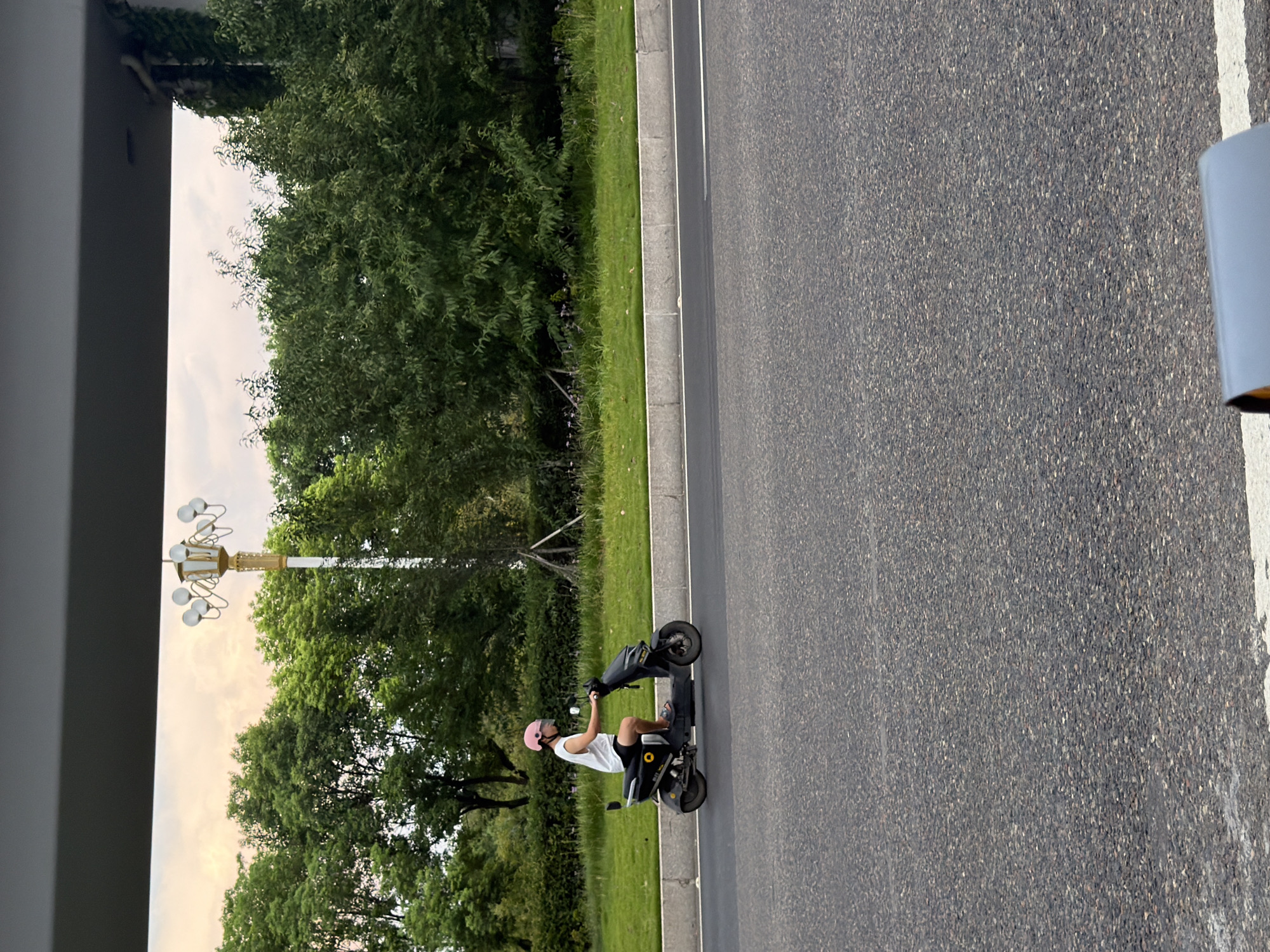 A person wearing a pink helmet and a white tank top with black shorts rides a black electric scooter on a paved road. The scooter has a yellow circular emblem on its side. The road is bordered by a concrete curb and a grassy area with trees and a lamppost in the background. The sky is overcast with a hint of sunset colors. The image appears to have been taken in the city of Jiangjin, China.